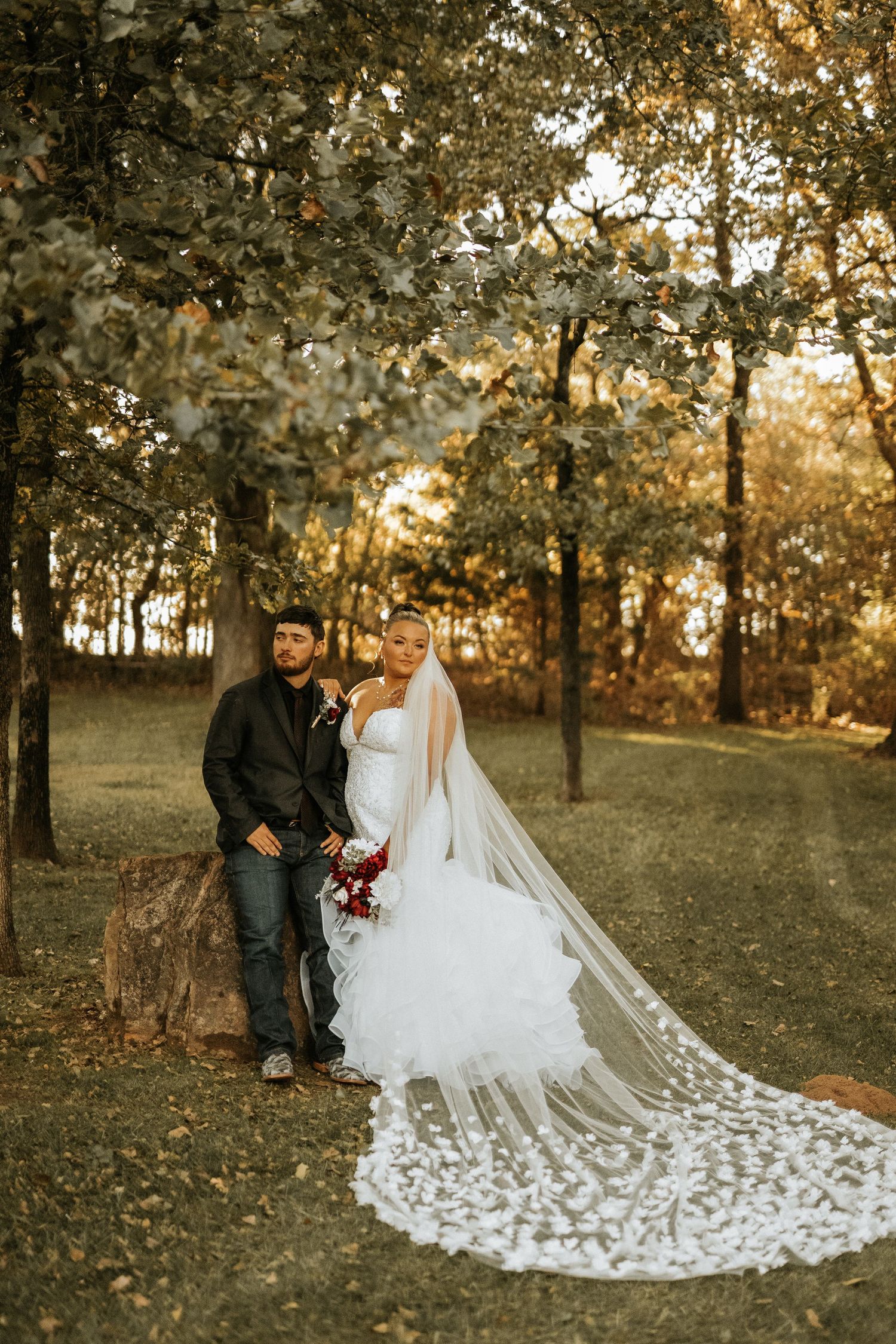 Bride in white gown with lace veil and groom in dark jacket pose together outdoors among autumn trees.