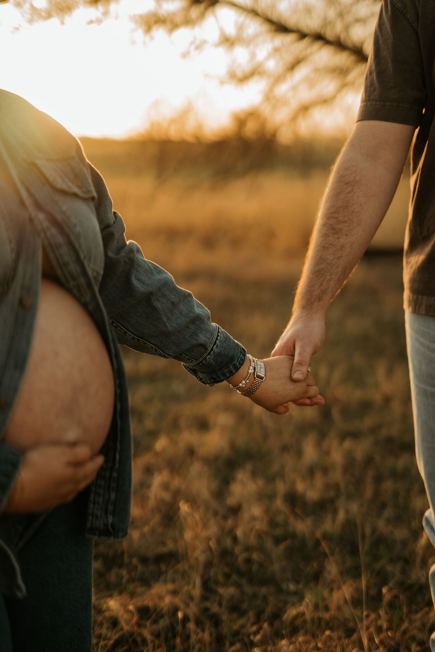 Pregnant woman and partner holding hands in golden sunset field, maternity photo shoot outdoors.