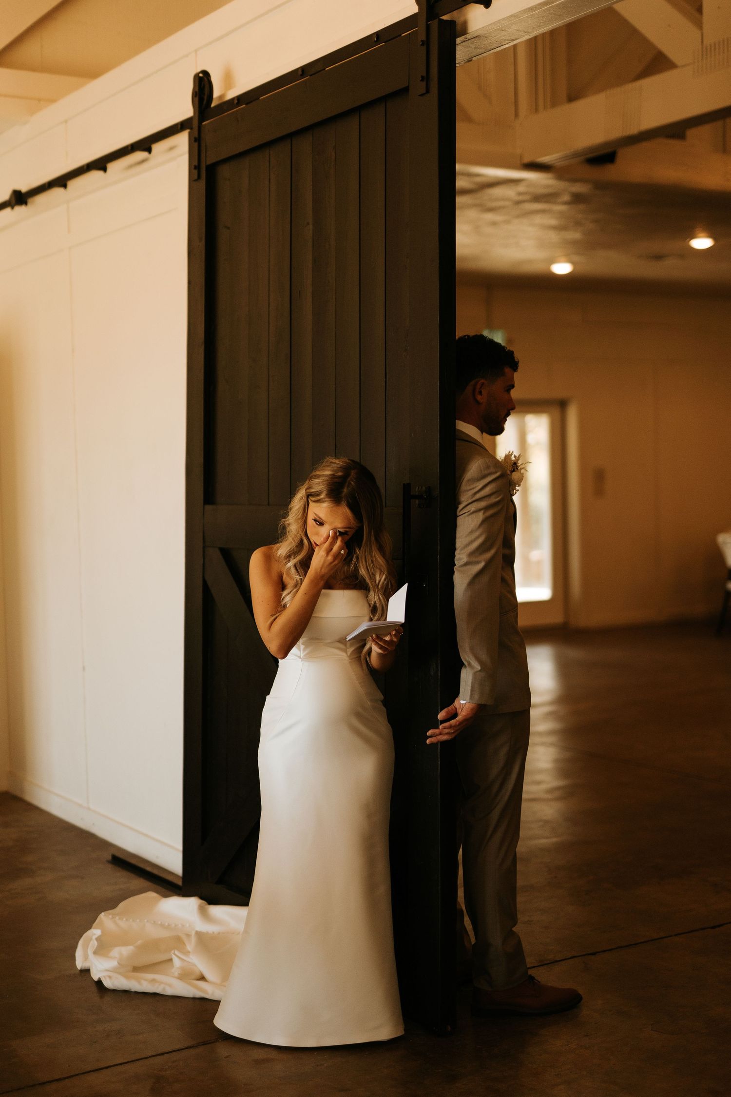 Bride in white gown reads vows while groom stands facing away at dark barn door during emotional first look.