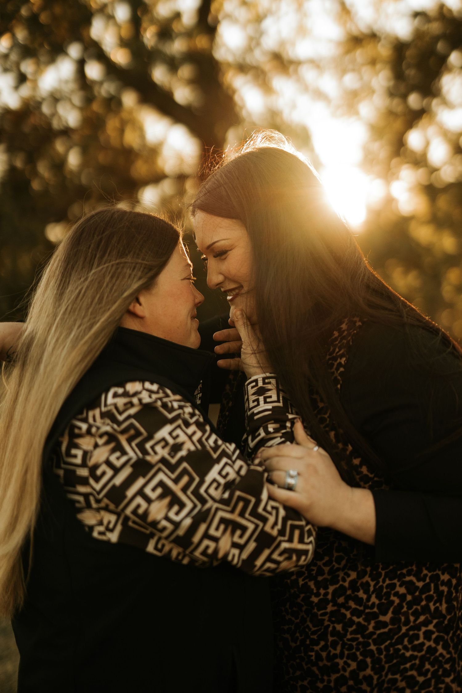 Two women share a tender moment at golden hour, smiling and embracing outdoors with warm sunlight glowing behind them.