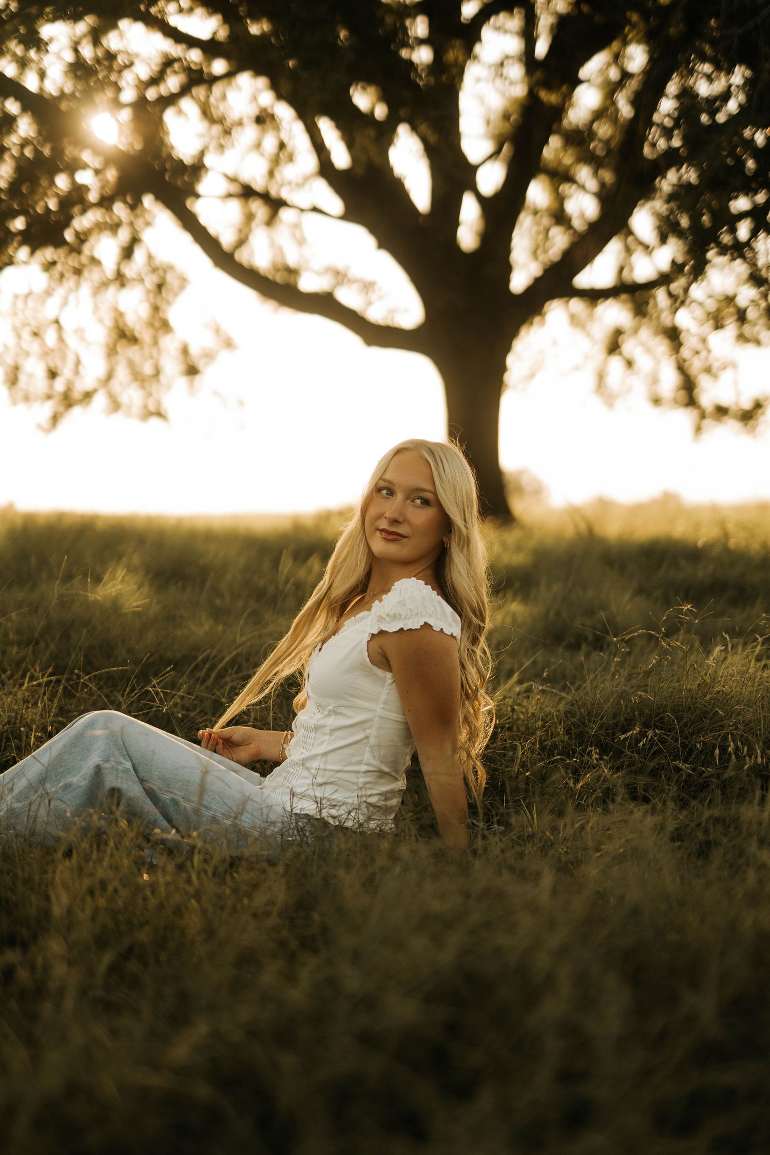 A young blonde woman sits in golden hour sunlight in a grassy field, wearing a white top and jeans, with a tree behind her.