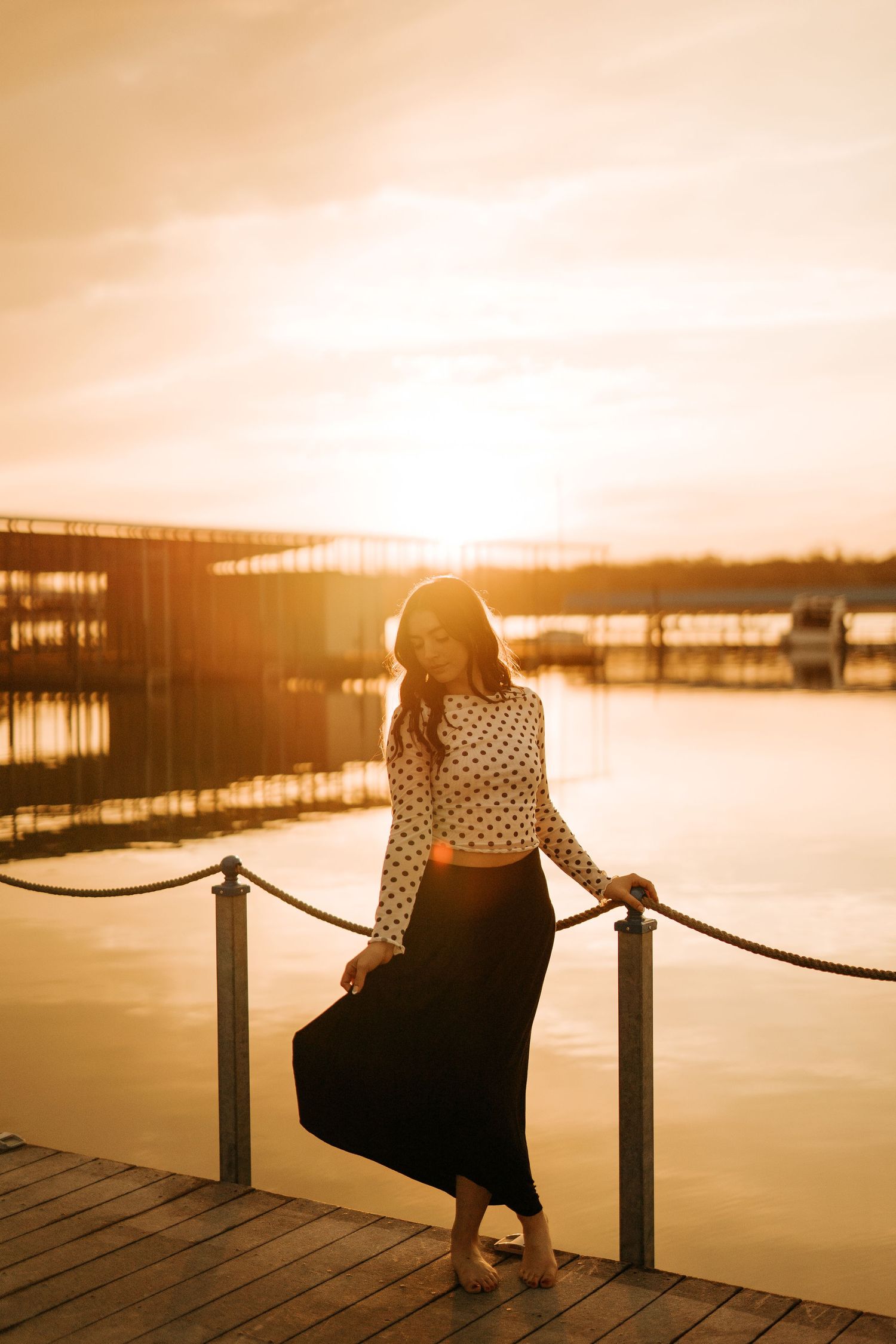 Woman in polka dot top and black skirt posing on a wooden dock at golden sunset with marina in background.