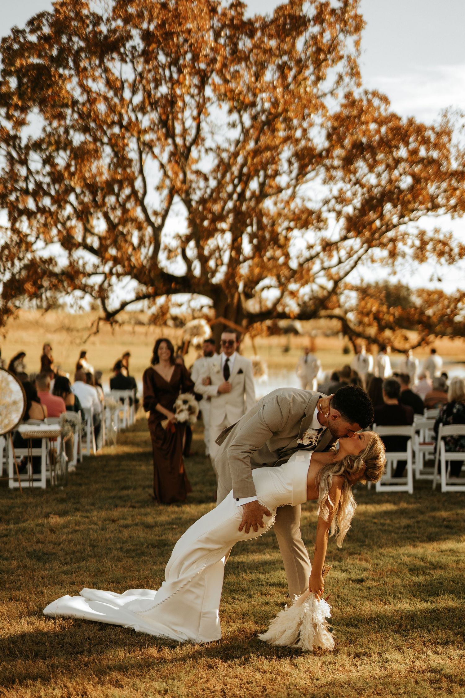 A groom dips his bride for a romantic kiss at an outdoor fall wedding ceremony under a golden oak tree.