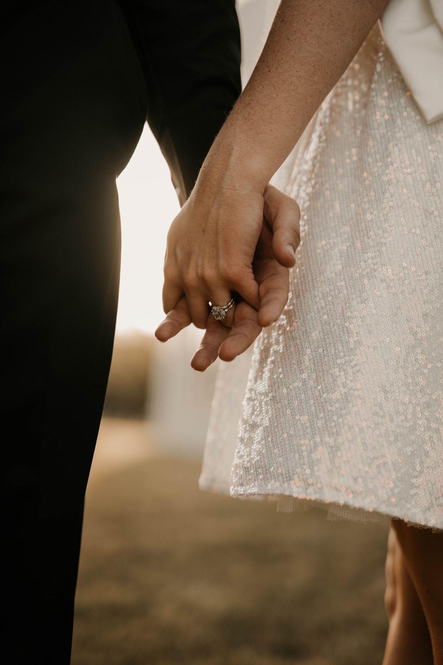 Close-up of newlyweds holding hands showing engagement ring and wedding attire details.