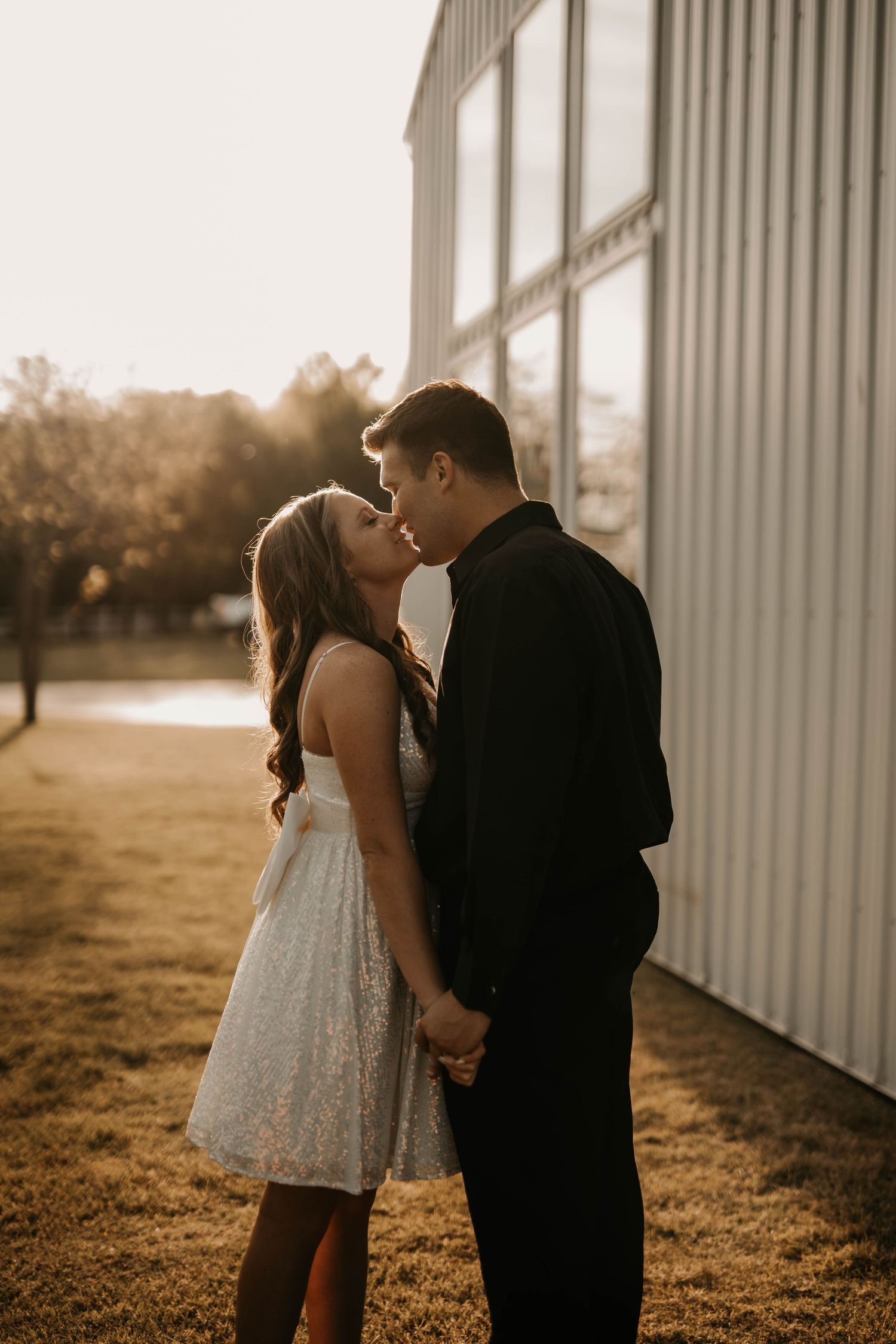 Couple in formal attire sharing an intimate moment against a metal building at sunset.