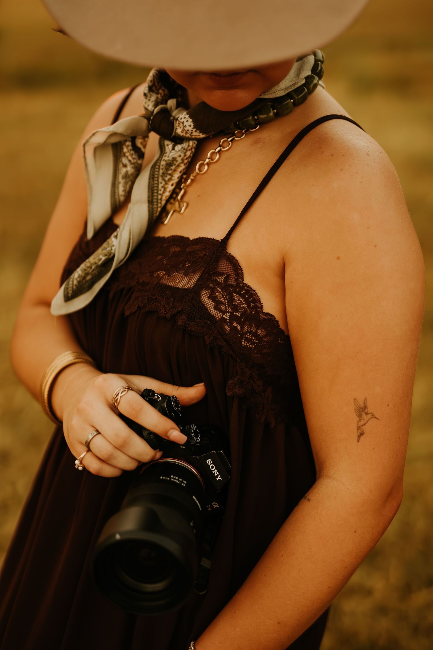 Woman in black lace dress and cowboy hat holding a Sony camera, wearing a silk scarf and gold necklace outdoors.