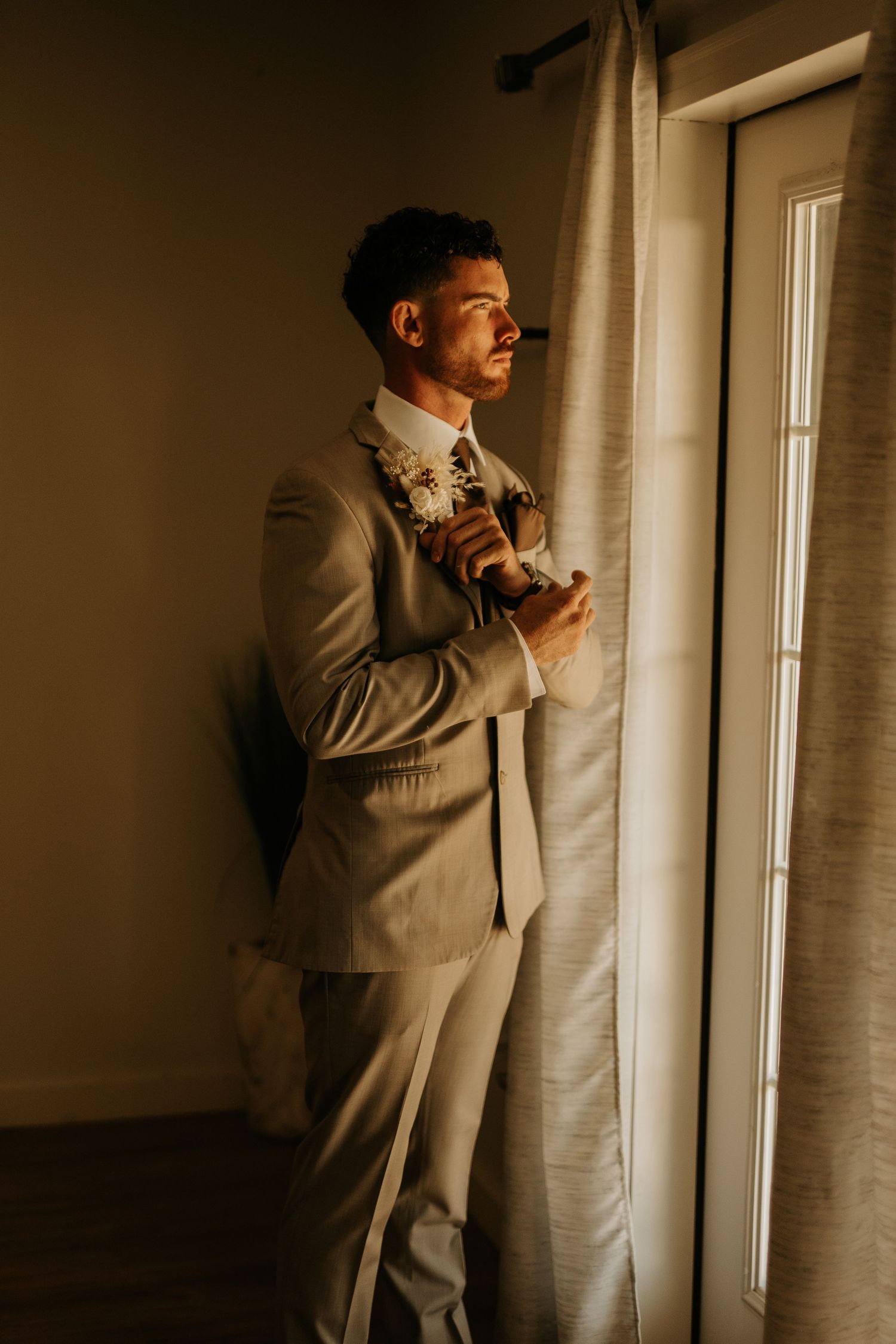 A groom in a beige suit with a floral boutonniere stands by a sunlit window, gazing thoughtfully outside.