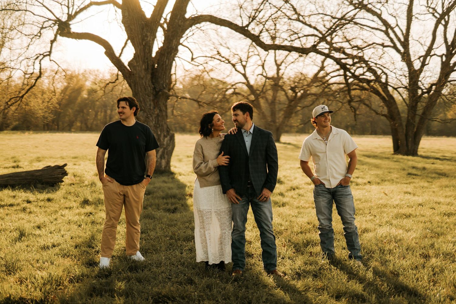 Four people standing in a sunlit field with bare trees, posing together during a golden hour outdoor photo session.
