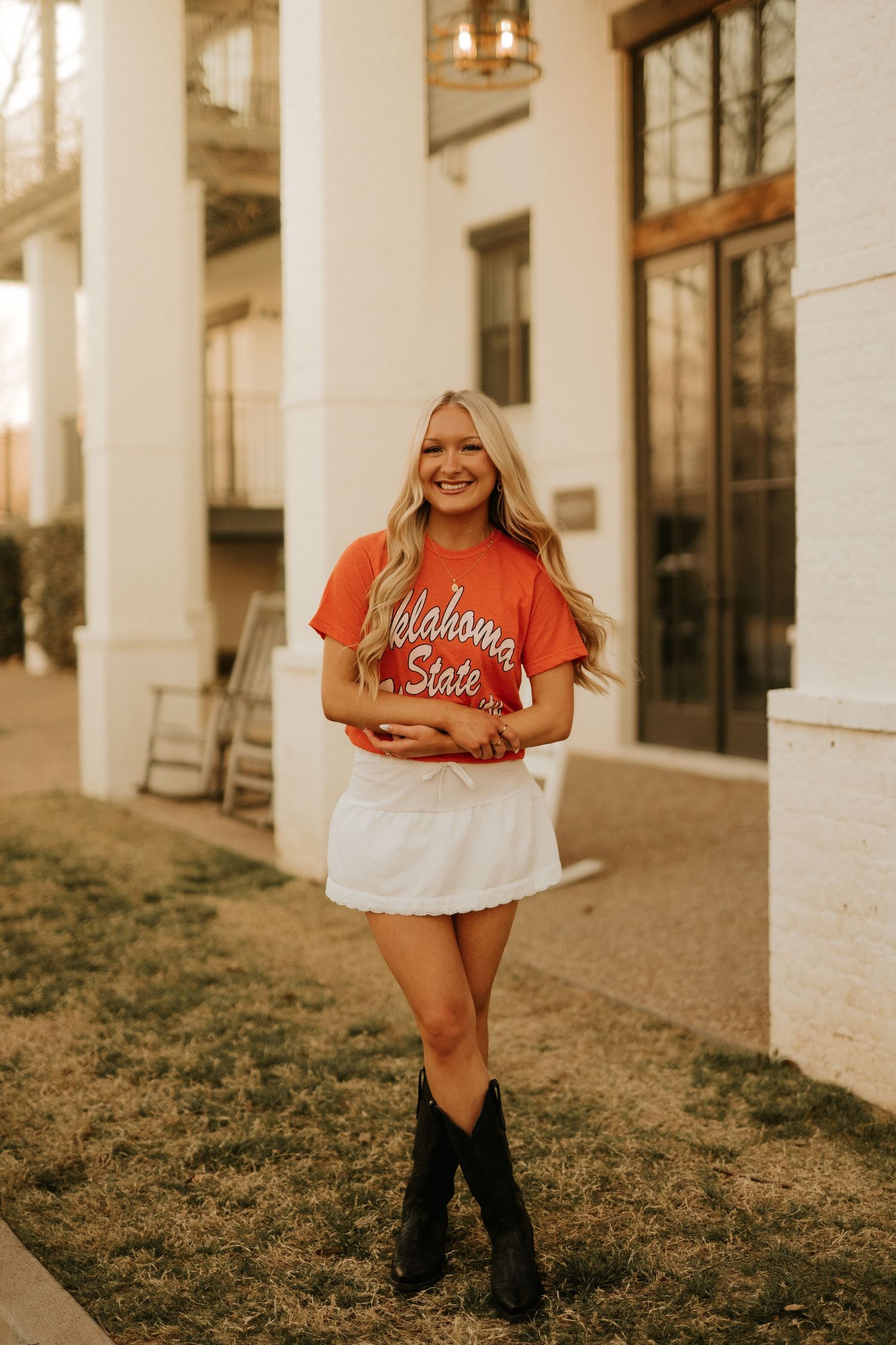 Smiling blonde woman wearing an Oklahoma State orange tee, white mini skirt, and black cowboy boots outdoors.
