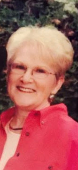Smiling elderly woman with short blonde hair and glasses wearing a red jacket outdoors.