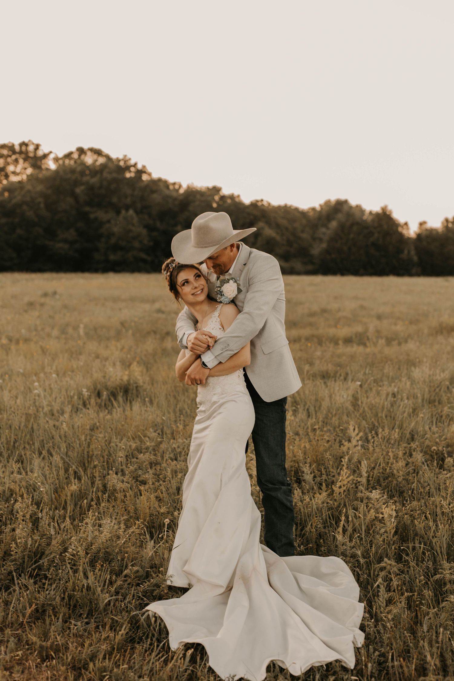 Romantic sunset portrait of couple embracing in field with flowing white dress and cowboy hat.