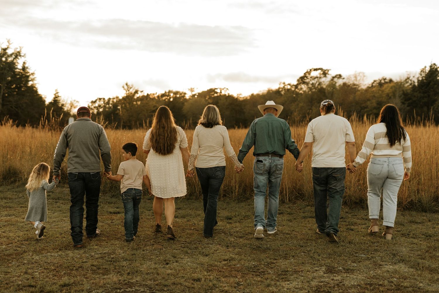 A multi-generational family holding hands walking through a golden field at sunset.