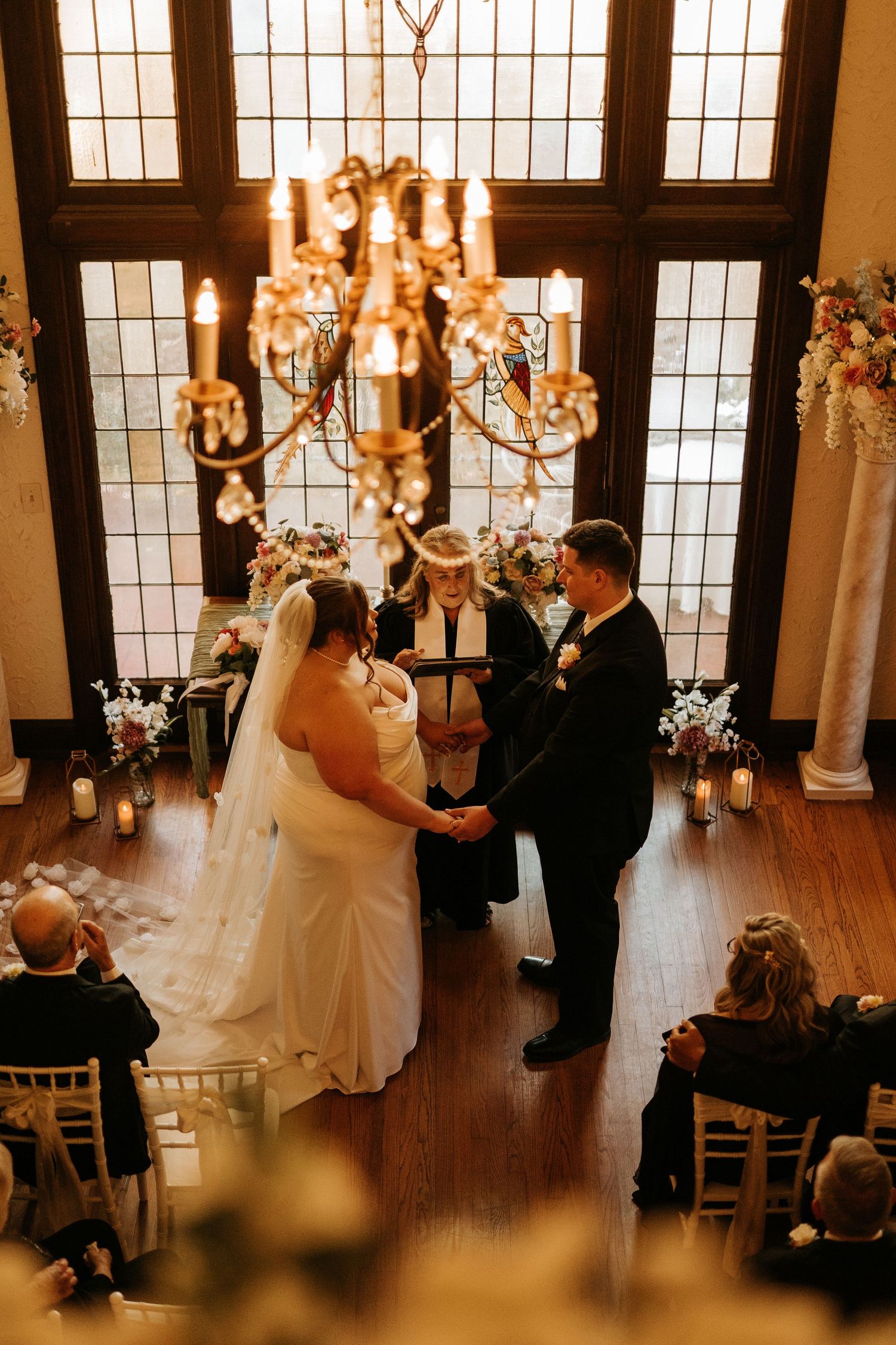 Bride and groom exchange vows beneath a glowing chandelier in an elegant indoor wedding ceremony with floral arrangements.