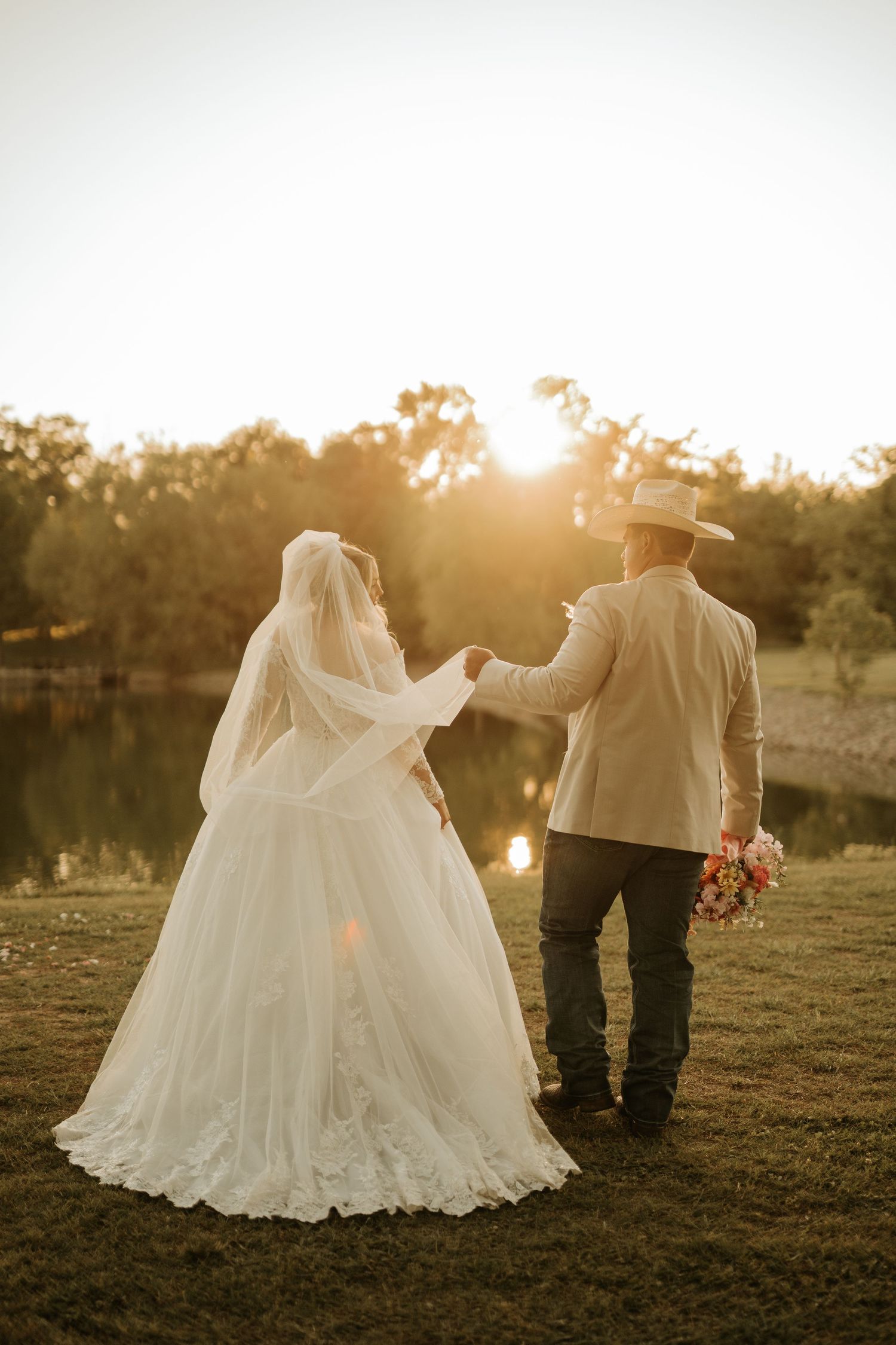 Bride and groom holding hands at golden sunset by a pond, bride in white gown with veil, groom in tan jacket and cowboy hat.