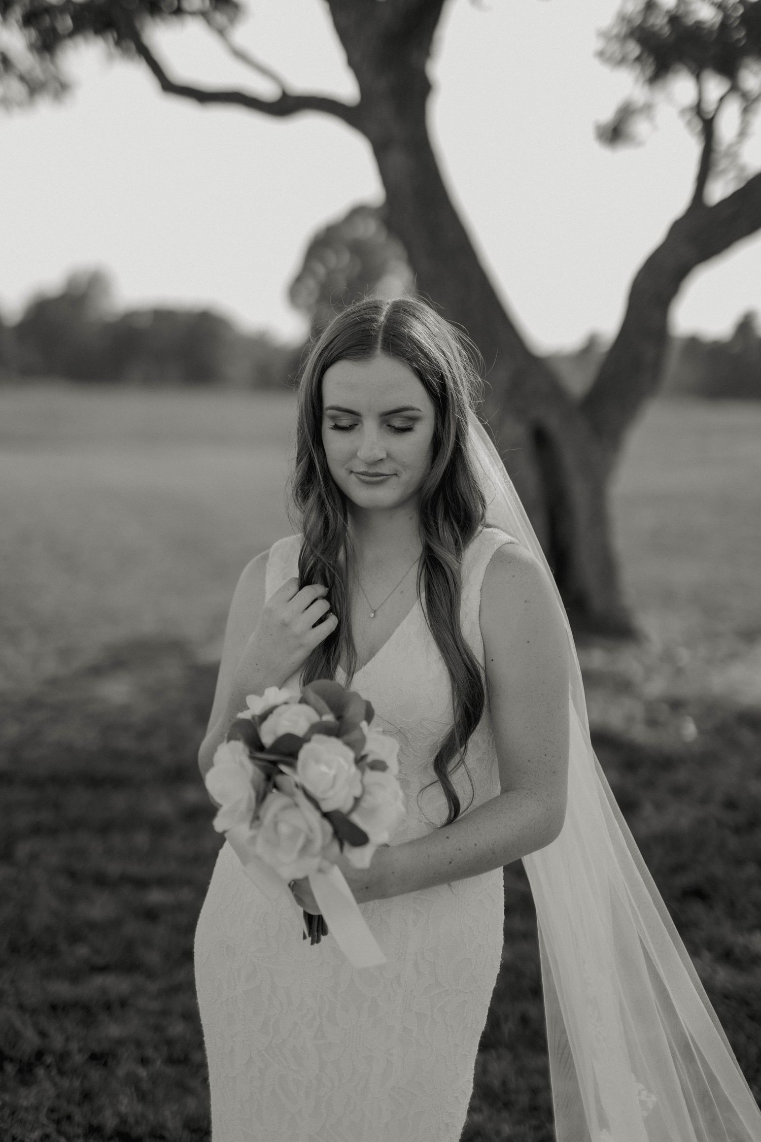 Bride in lace dress with long veil holds bouquet while looking down during outdoor bridal portrait session at sunset.