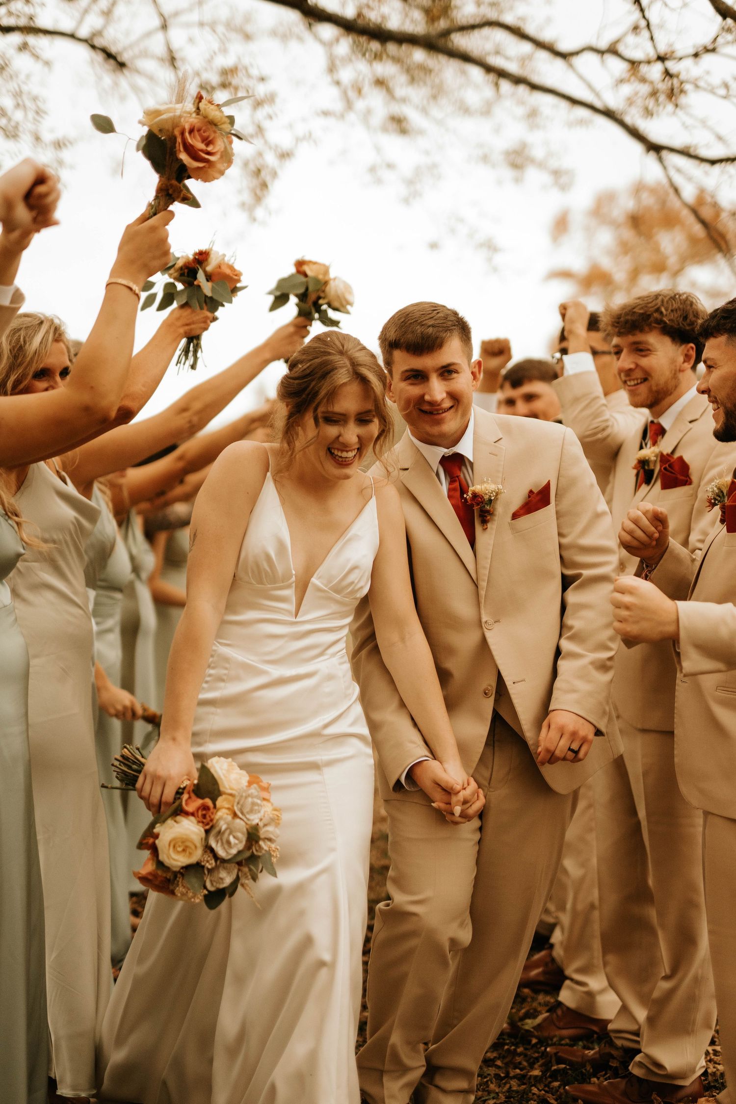 Wedding party celebrates with raised bouquets as newlyweds walk through in tan and white attire.