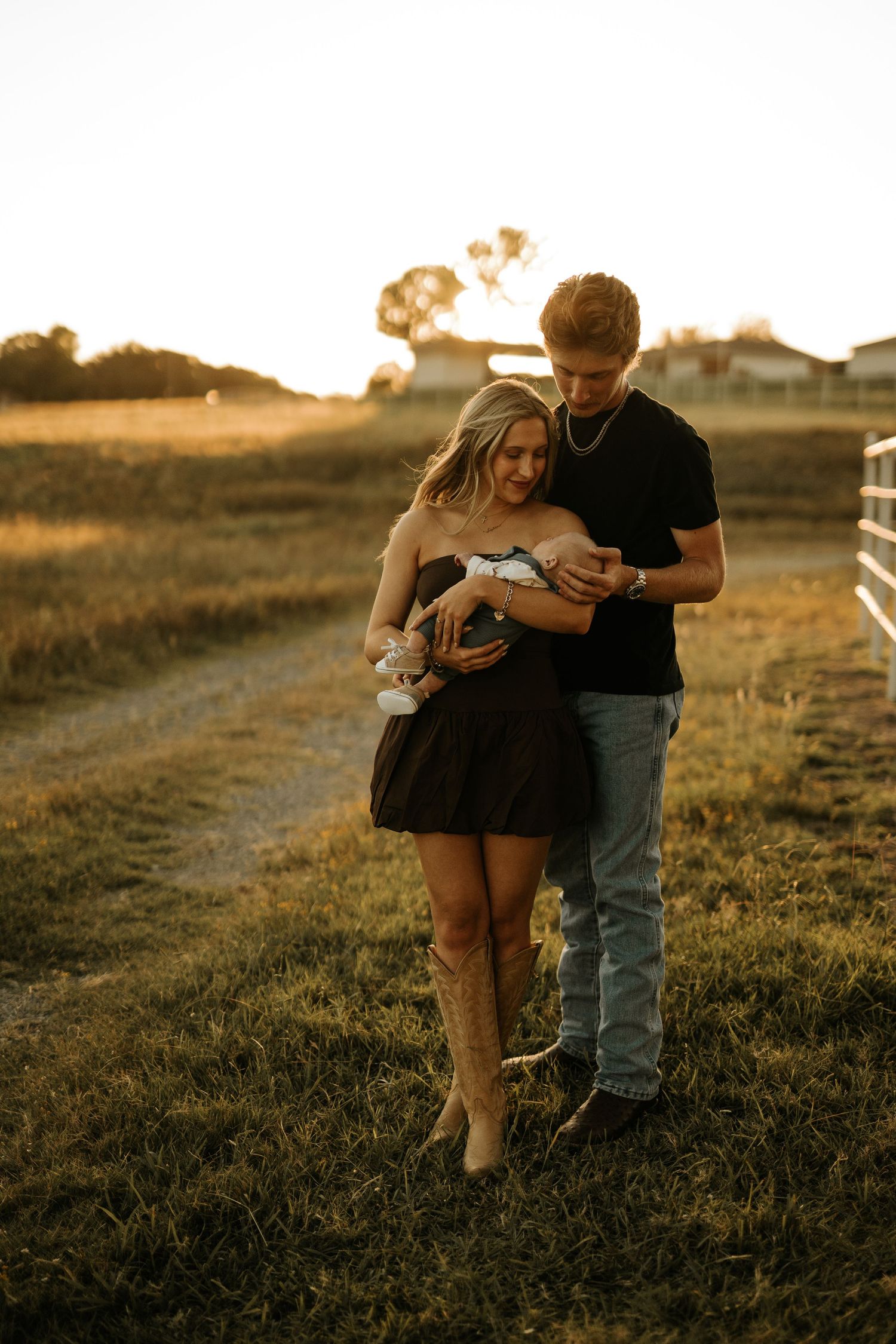 Young couple in golden hour light holding newborn baby on rural farm path, woman in black dress and cowboy boots.