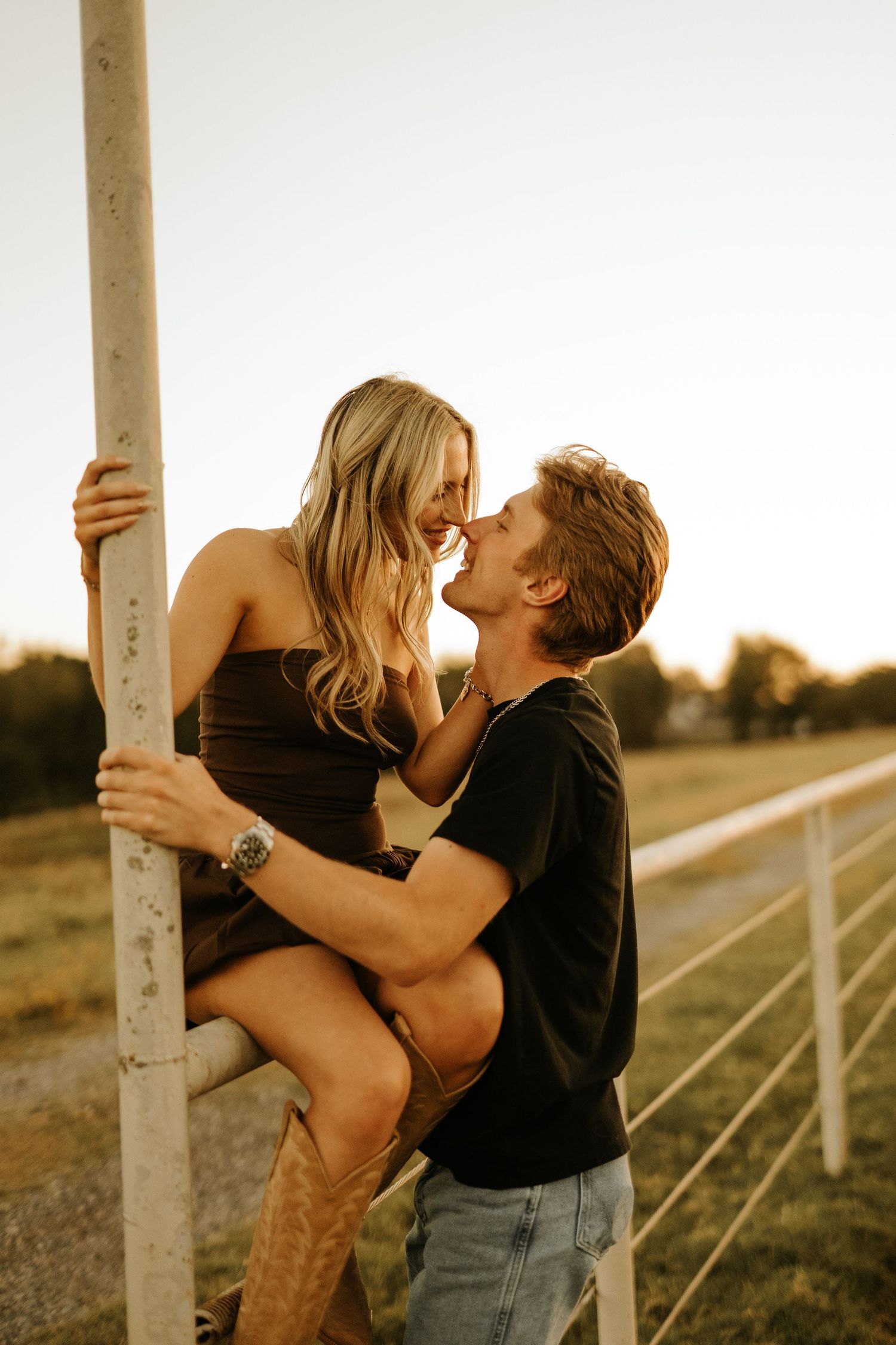 Couple sharing a romantic moment near a fence post at golden hour on a farm, woman in brown dress and cowboy boots.