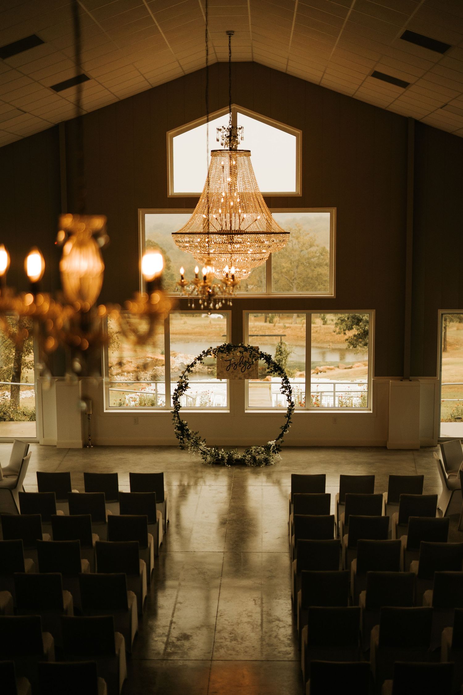 Elegant wedding venue interior with crystal chandeliers, floral hoop arch, and rows of dark chairs under vaulted ceilings.