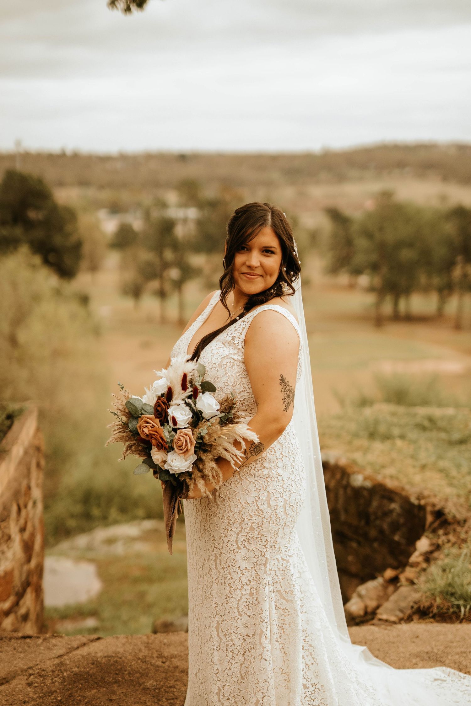 Bride in lace gown holds autumn-colored bouquet against rolling countryside backdrop.