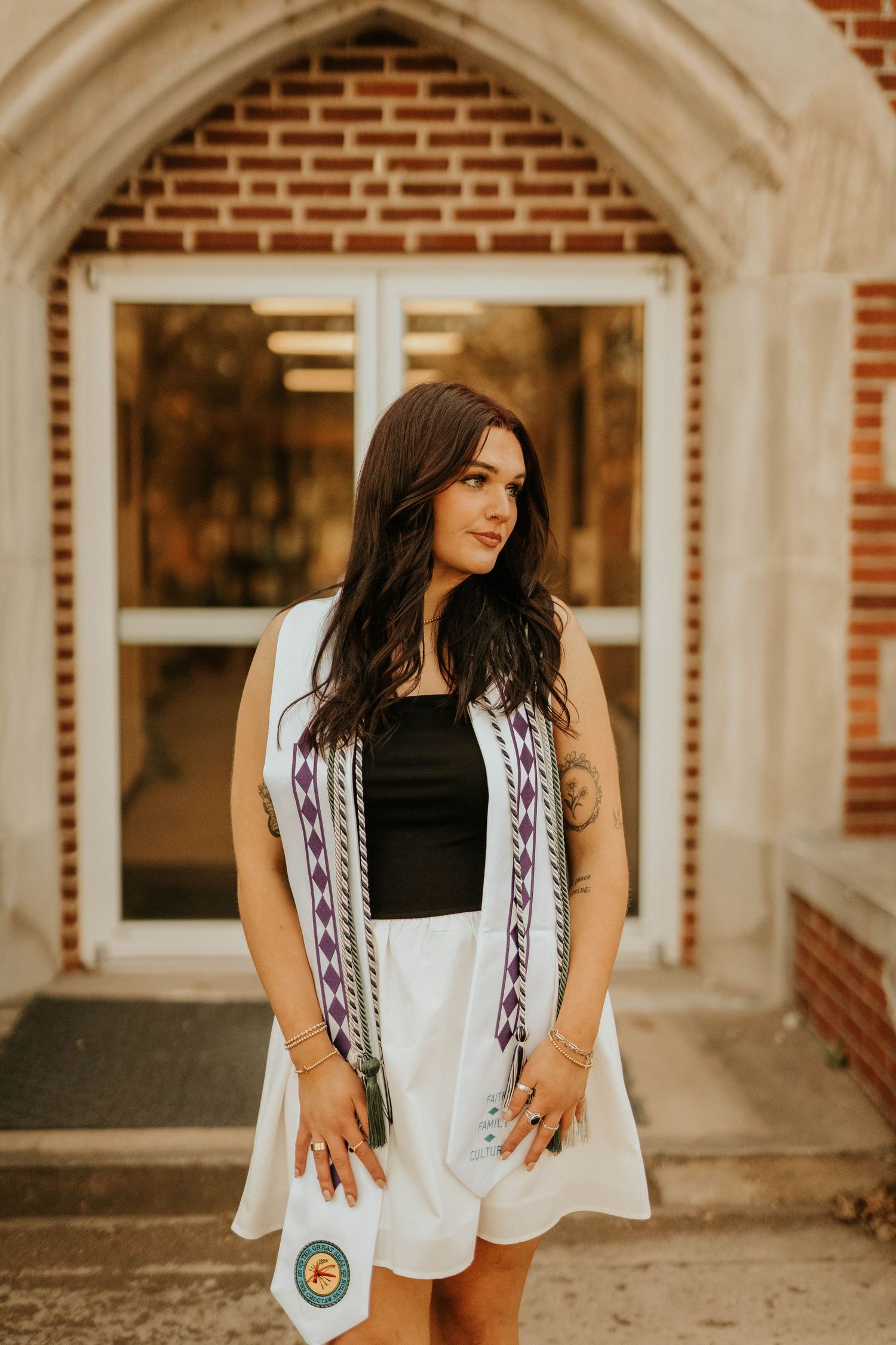 Graduate wearing white dress and patterned stole stands in front of arched doorway.