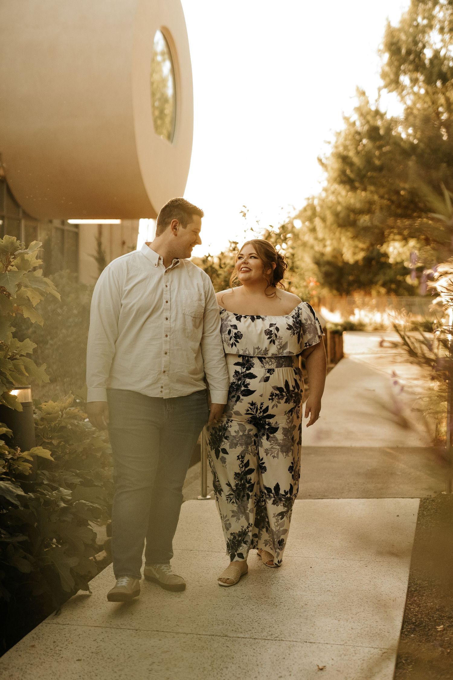 Couple walking hand in hand on a sunlit path, woman in floral jumpsuit and man in light shirt, sharing a joyful smile.
