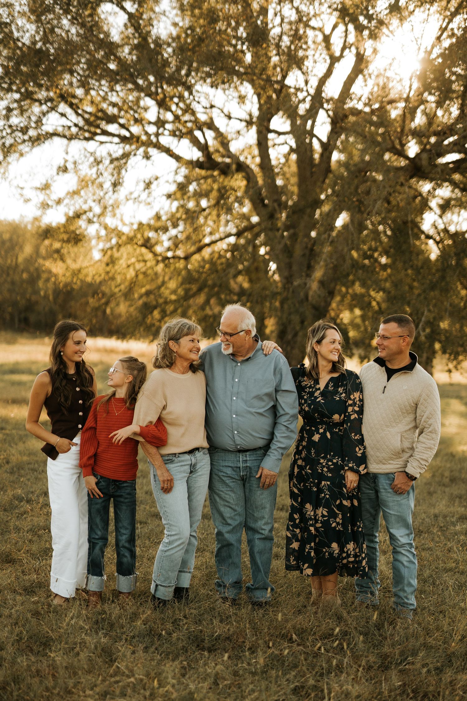 Multi-generational family of six smiling together in a golden sunlit field with large oak trees in the background.