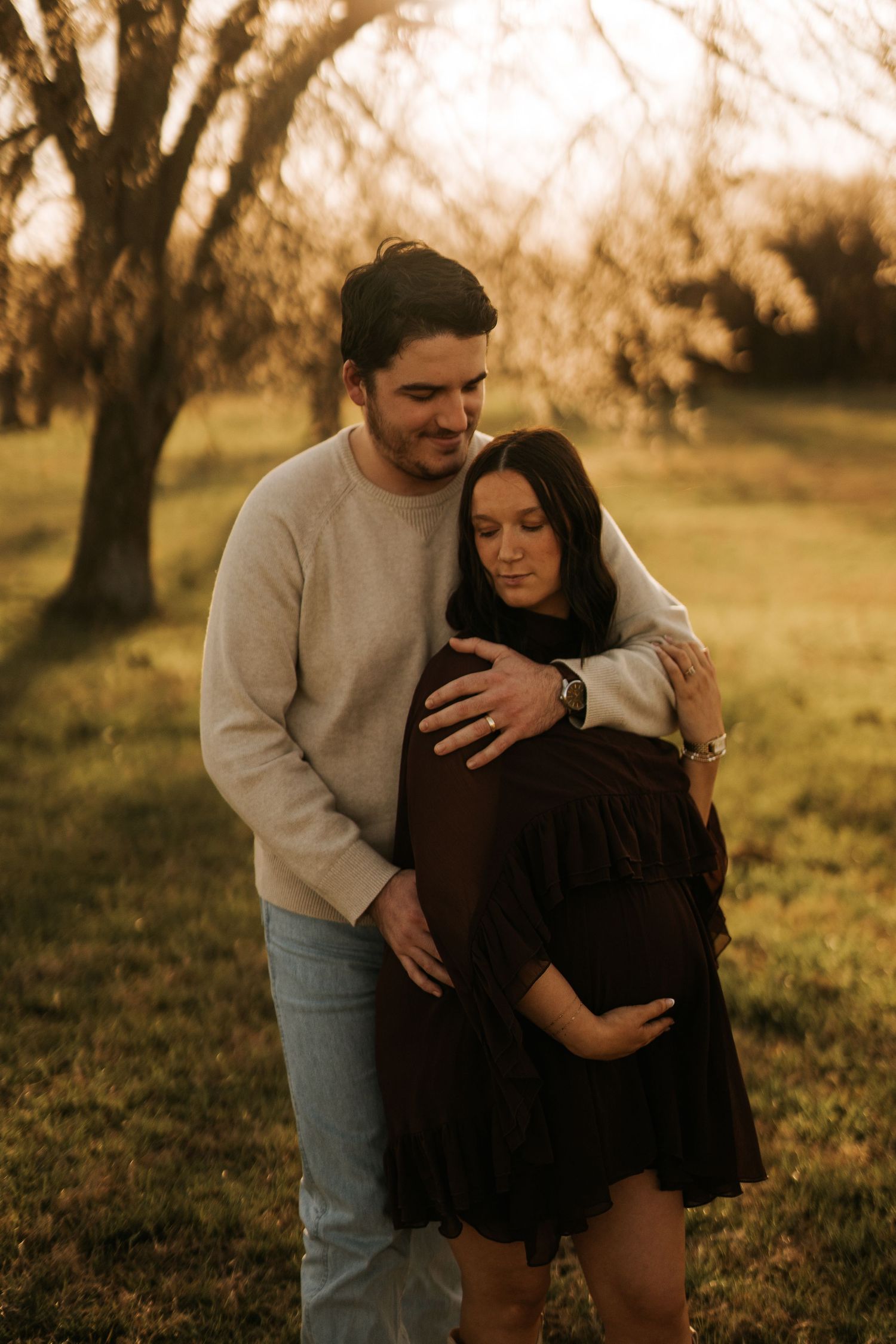 Couple sharing a tender maternity moment outdoors at golden hour, man embracing pregnant woman in dark dress.