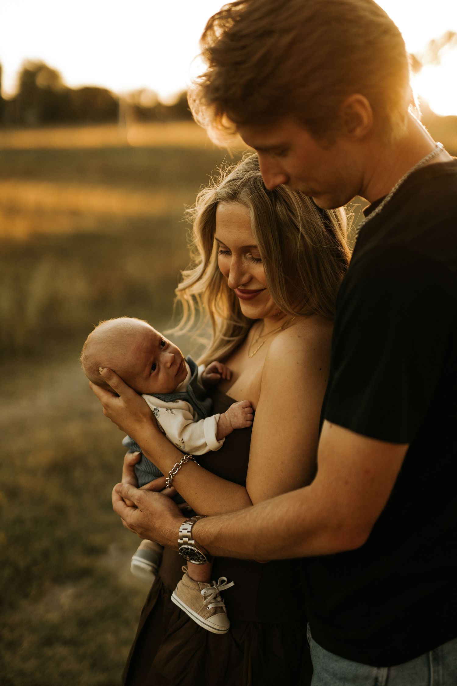 A young couple shares a tender moment outdoors at golden hour, lovingly cradling their newborn baby in a sunlit field.