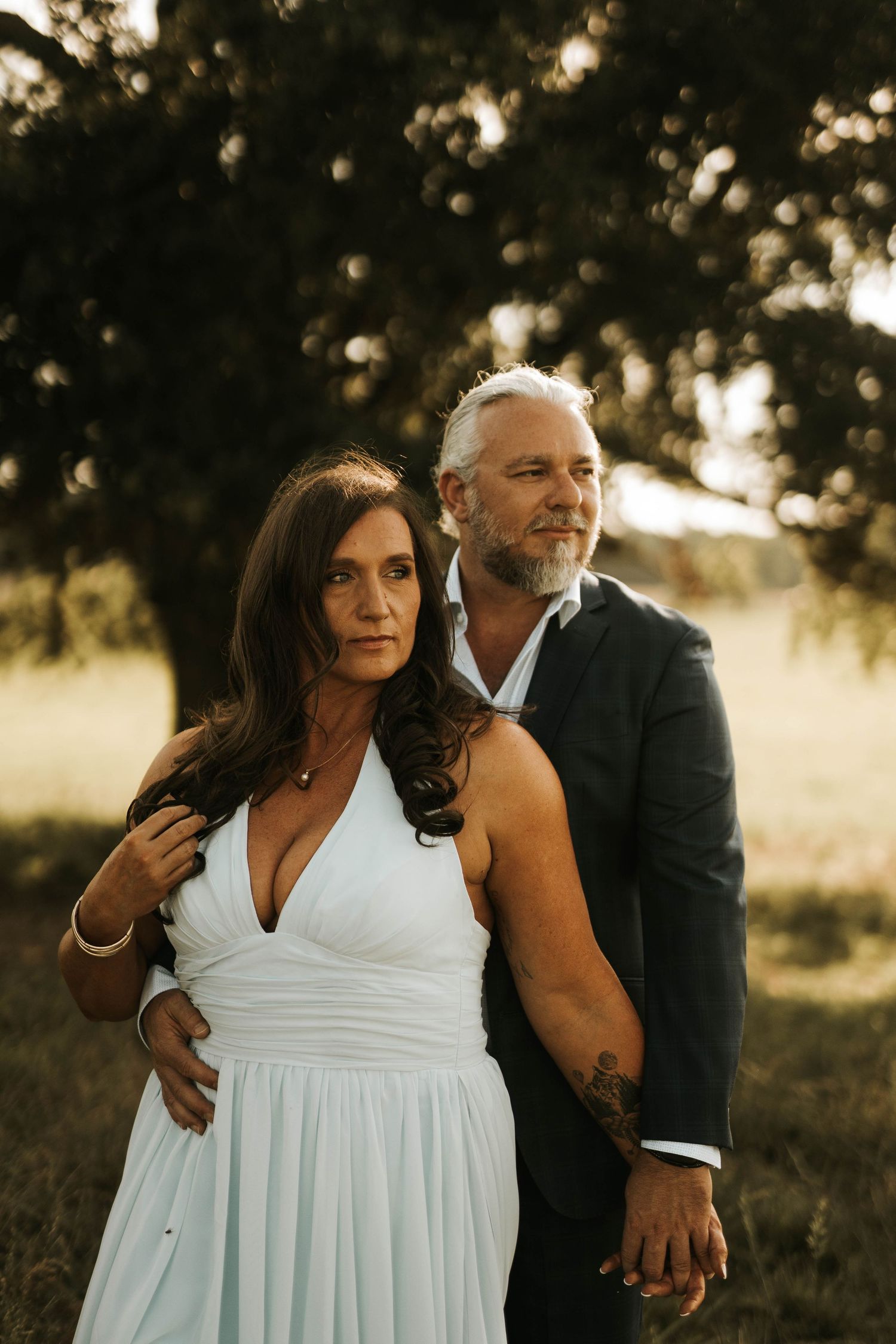 A couple in formal attire poses outdoors at golden hour, woman in white dress, man in dark suit with beard and tattoo.