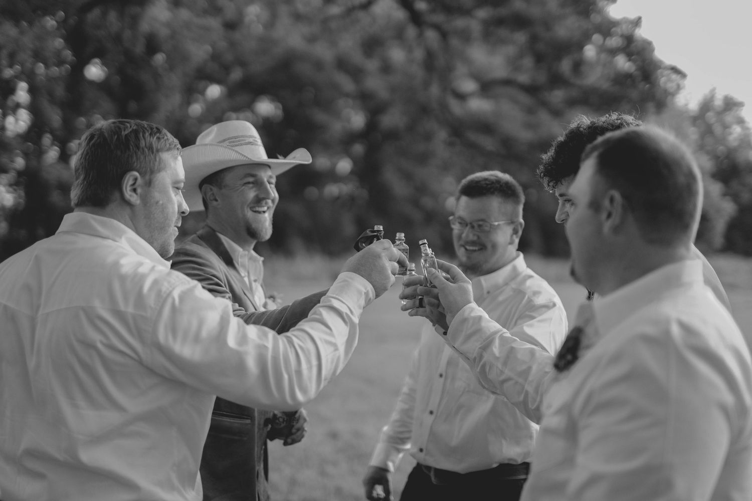 Black and white photo of groomsmen in white shirts toasting drinks outdoors, one wearing a cowboy hat at a wedding.