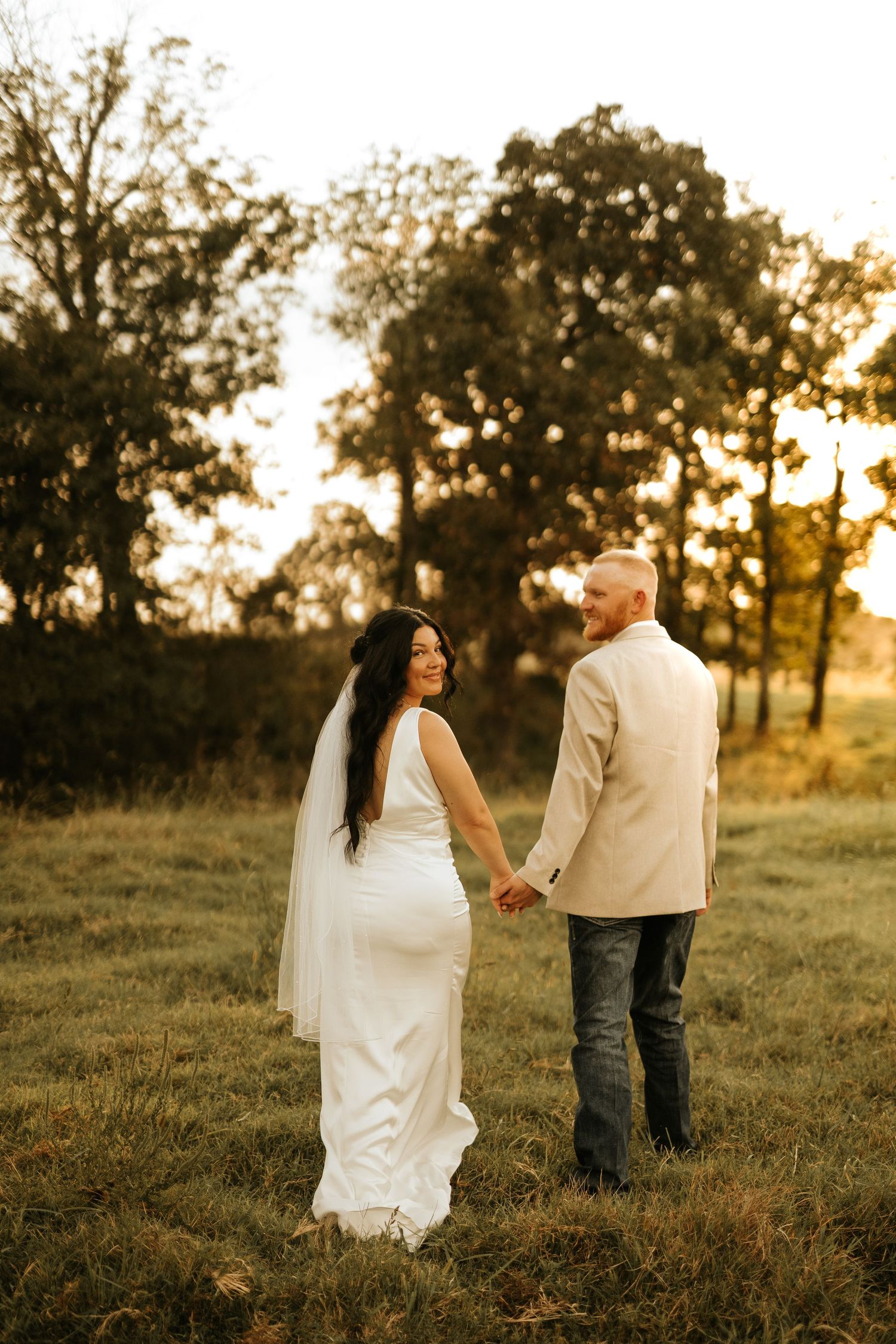 A bride and groom hold hands in a golden sunset field, she smiles back in a white gown and veil, he wears a tan suit.