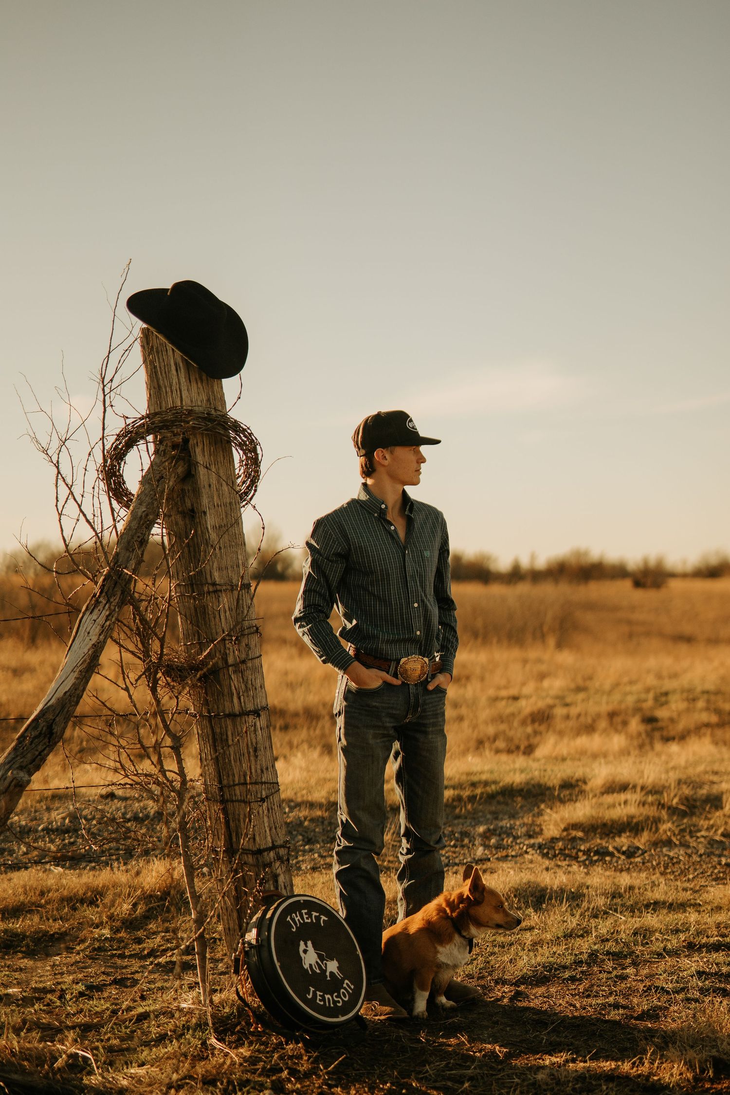 A cowboy stands in a golden field beside a wooden fence post with a black hat, accompanied by a corgi dog at sunset.