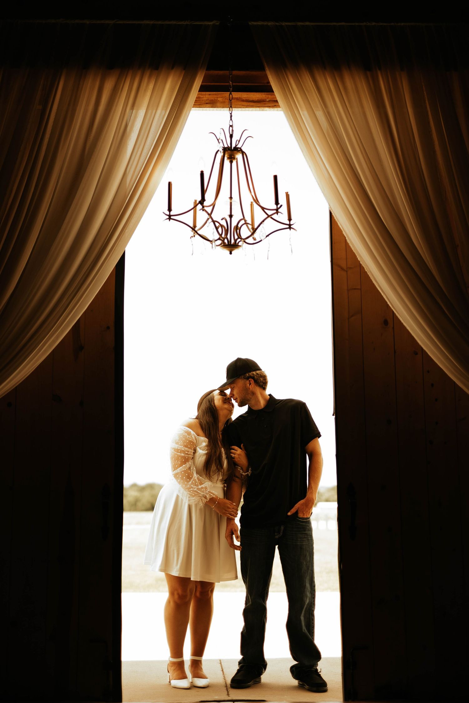 Silhouetted couple poses in front of window with elegant chandelier and flowing curtains.