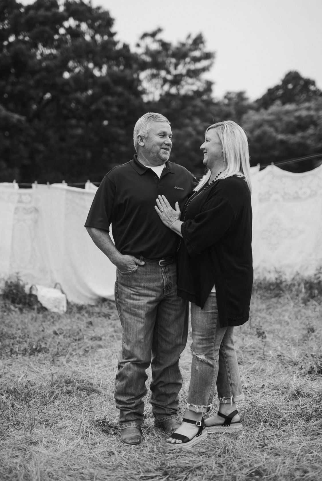 Black and white photo of a smiling couple standing together outdoors near hanging white linens and trees.