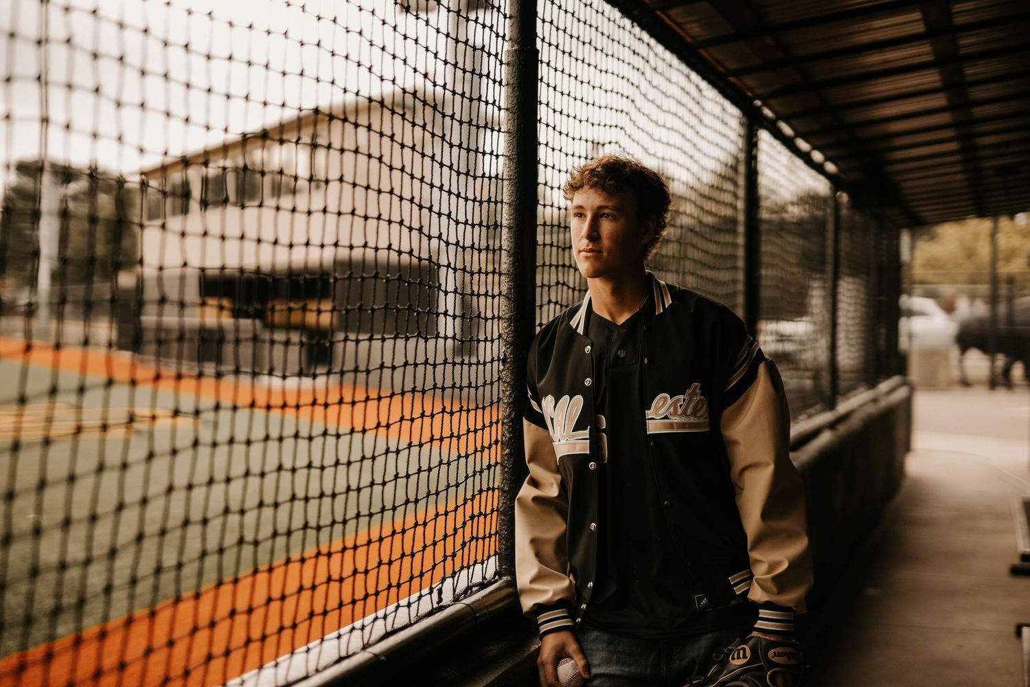 Athletic figure stands by mesh fence in warm sepia lighting wearing varsity jacket and casual attire.
