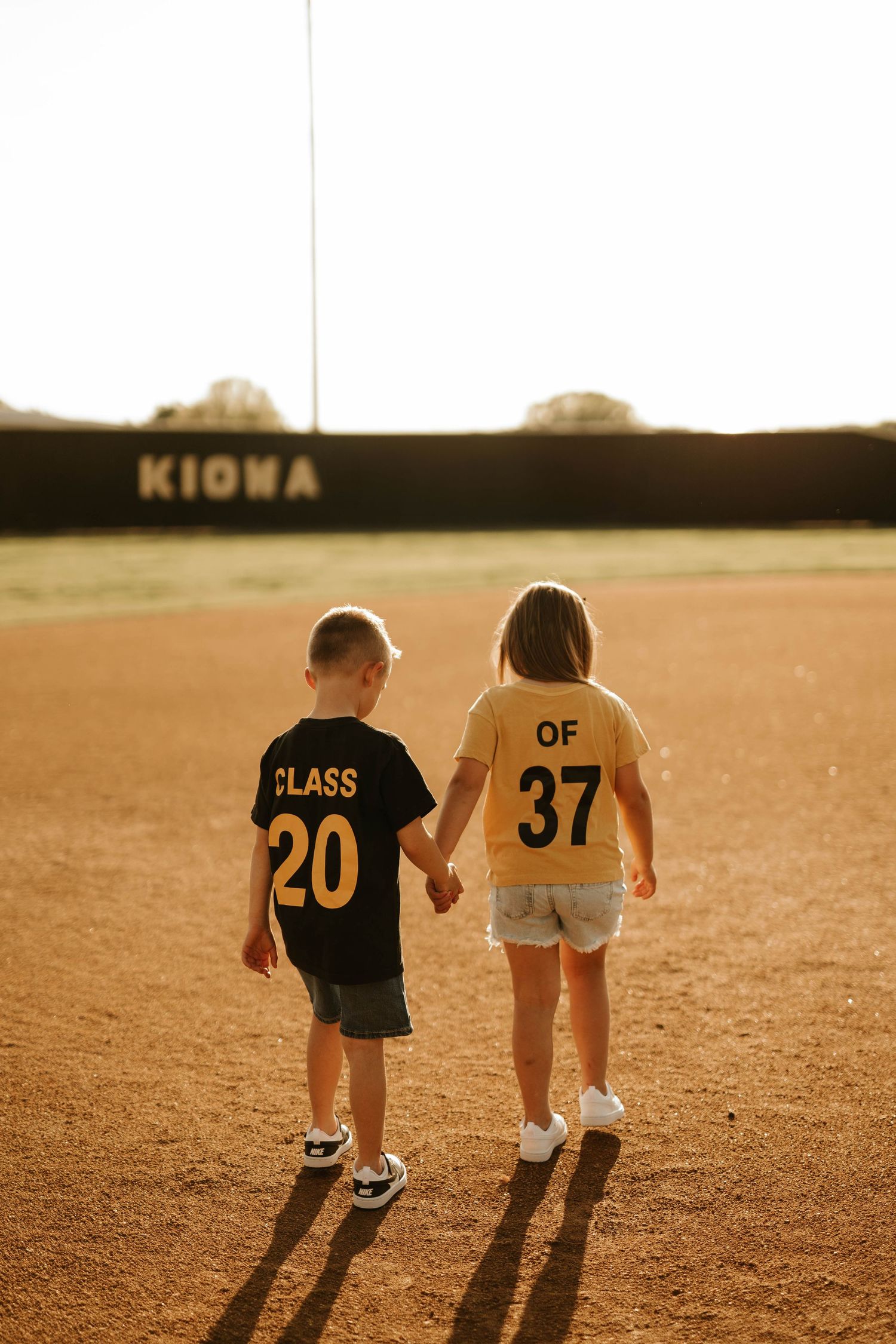Two children wearing sports jerseys walking on baseball field at sunset.