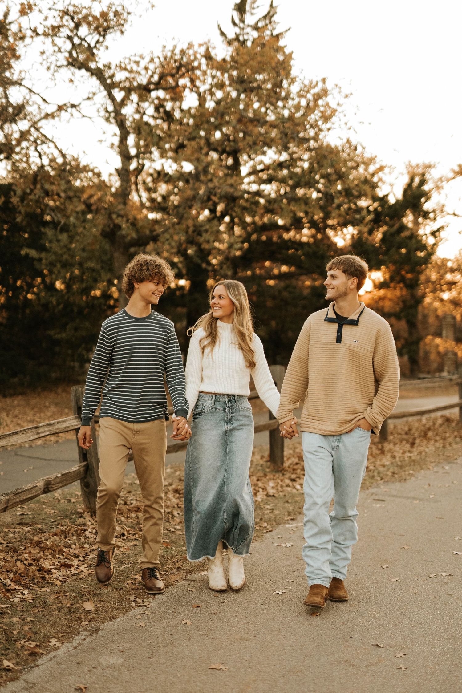 Three friends holding hands while walking on autumn path surrounded by golden trees.