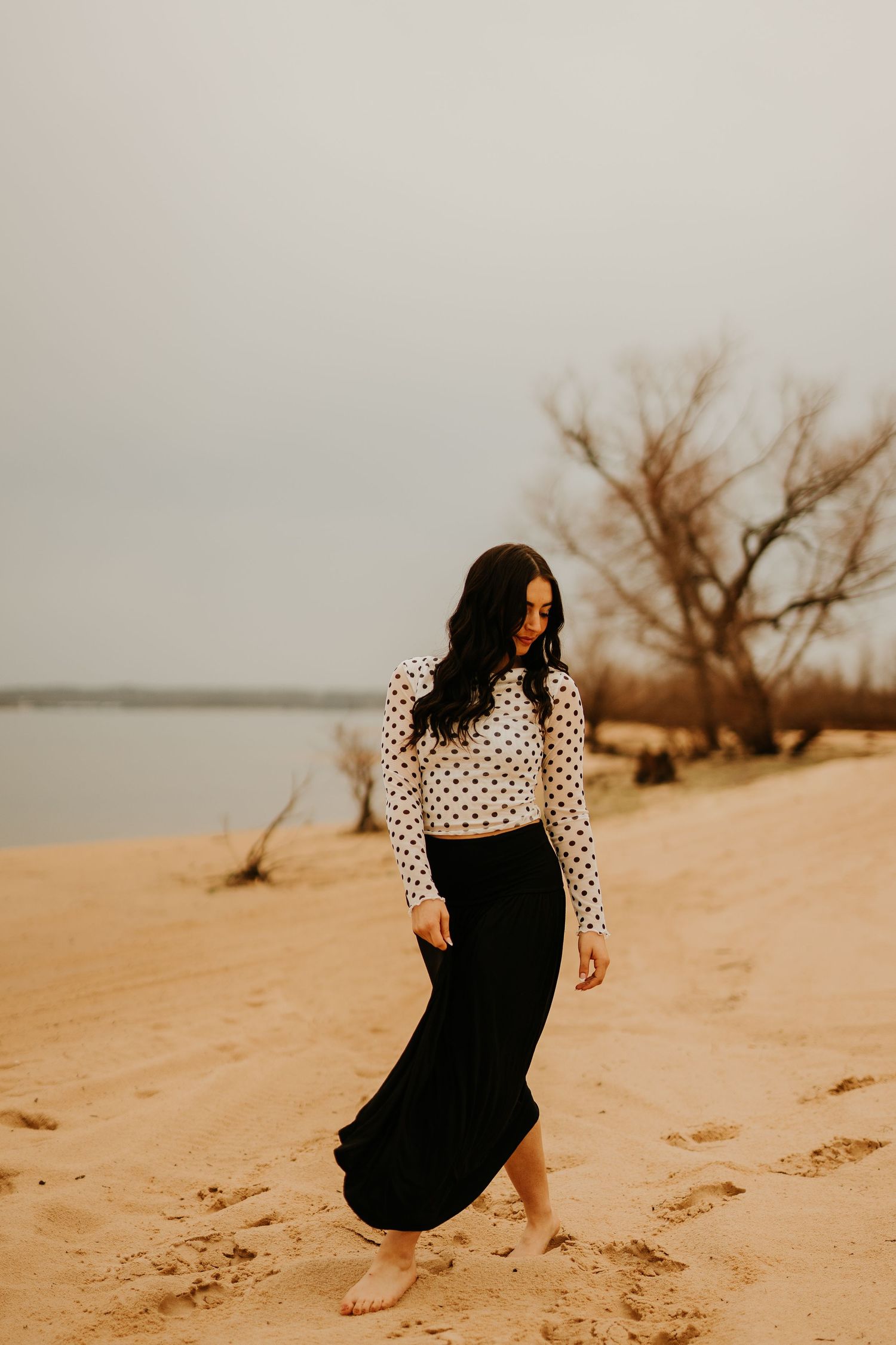 Woman in polka dot top and black maxi skirt walking barefoot on sandy beach with bare trees and water in background.