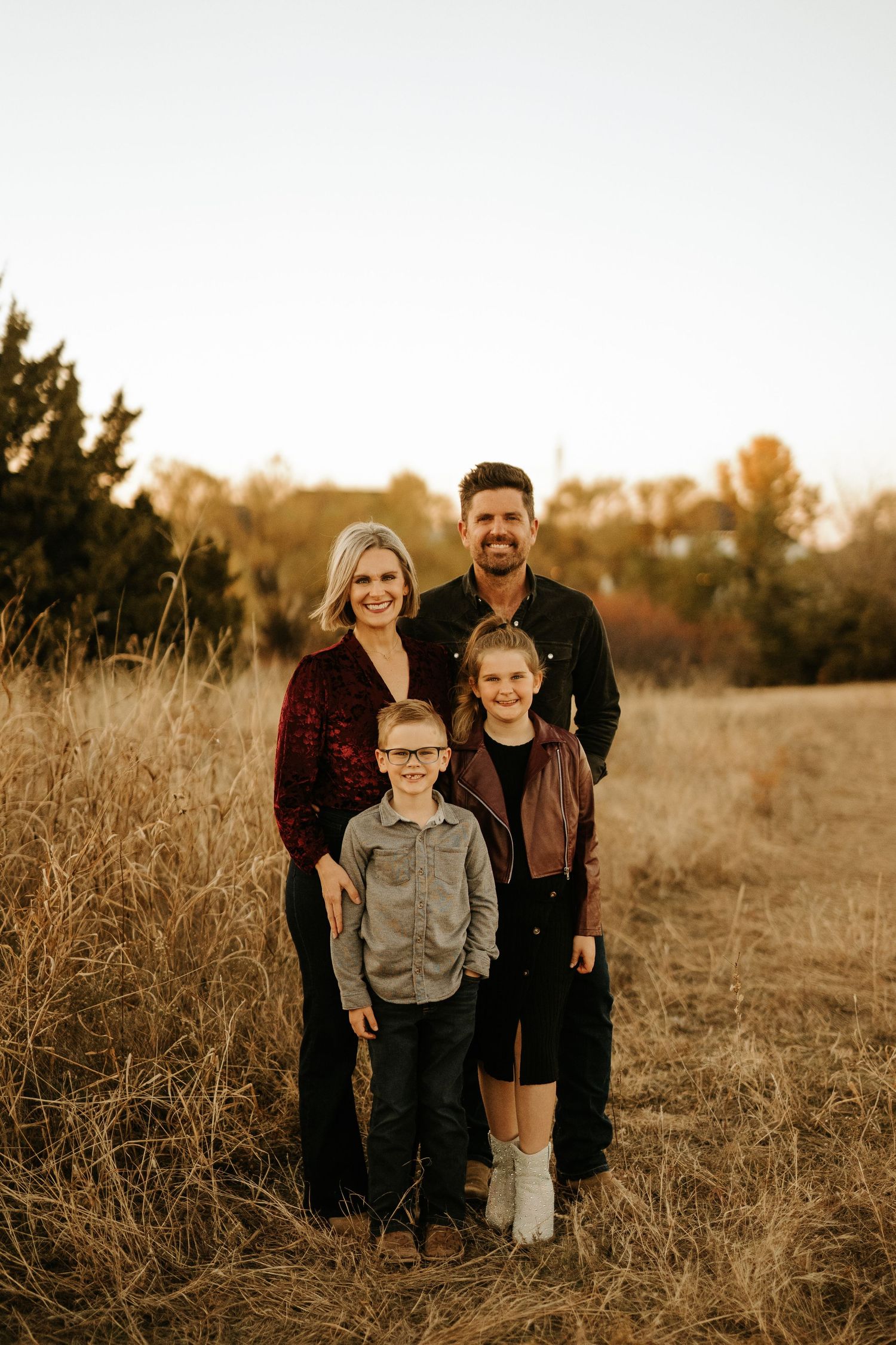 Family of four posing in a golden autumn field, smiling outdoors during fall season photo shoot.