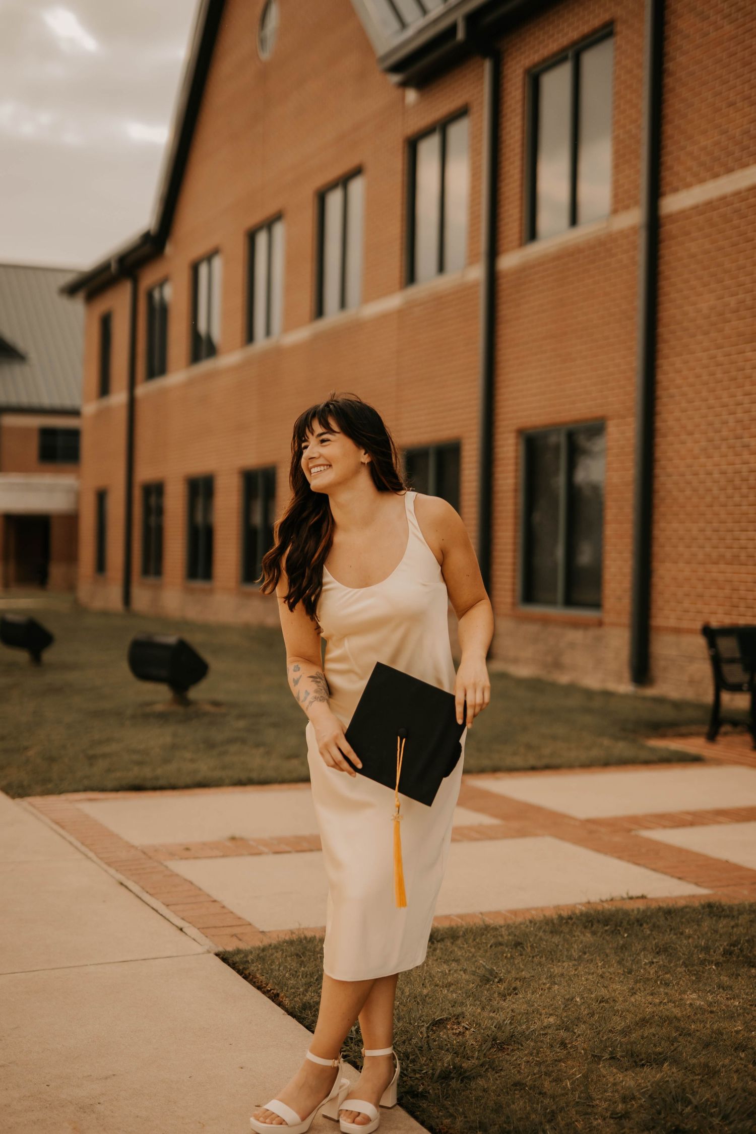 Person in cream dress holds diploma while standing outside brick building.