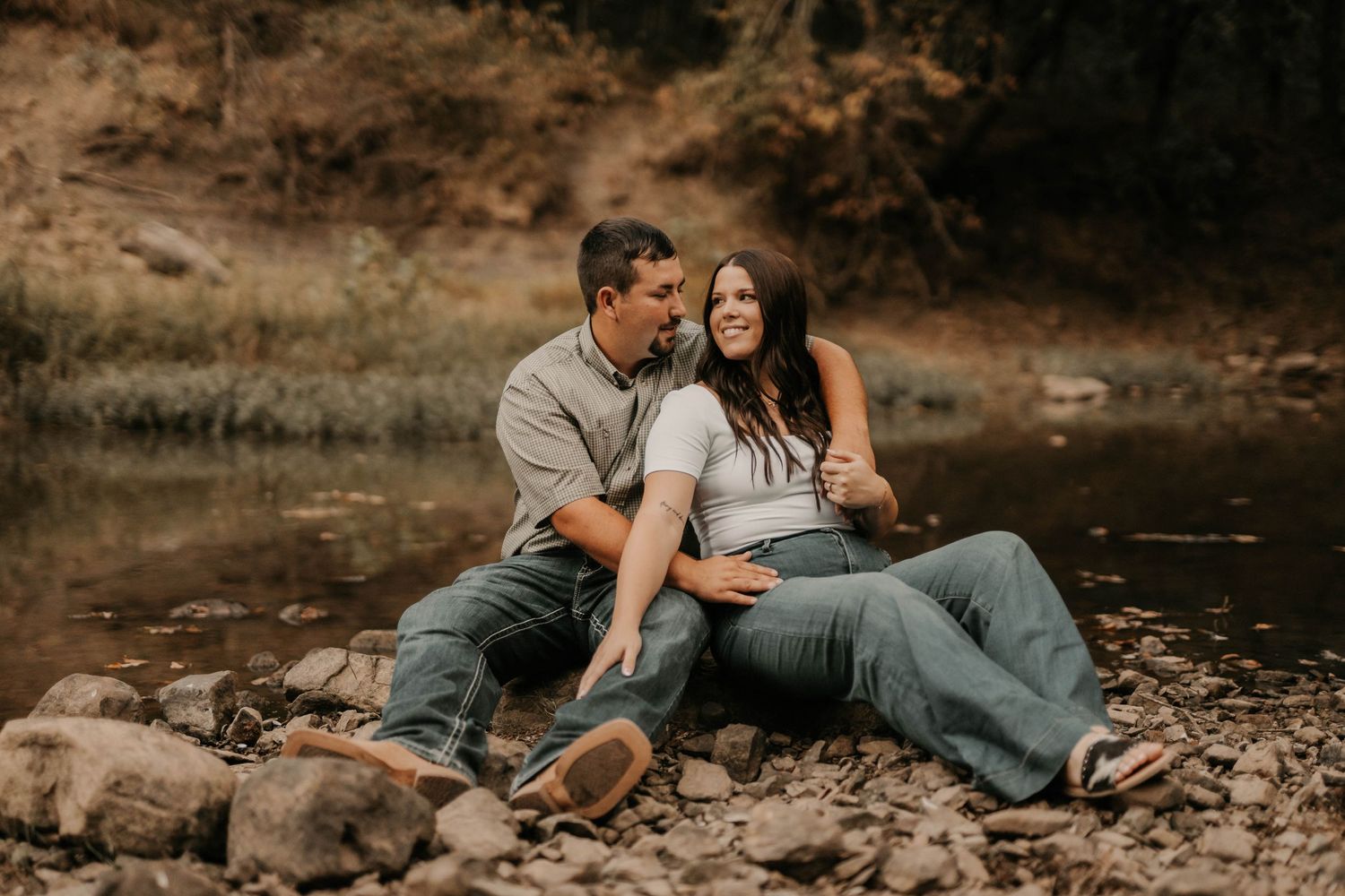 Couple relaxing together on rocky riverbank during romantic photoshoot.