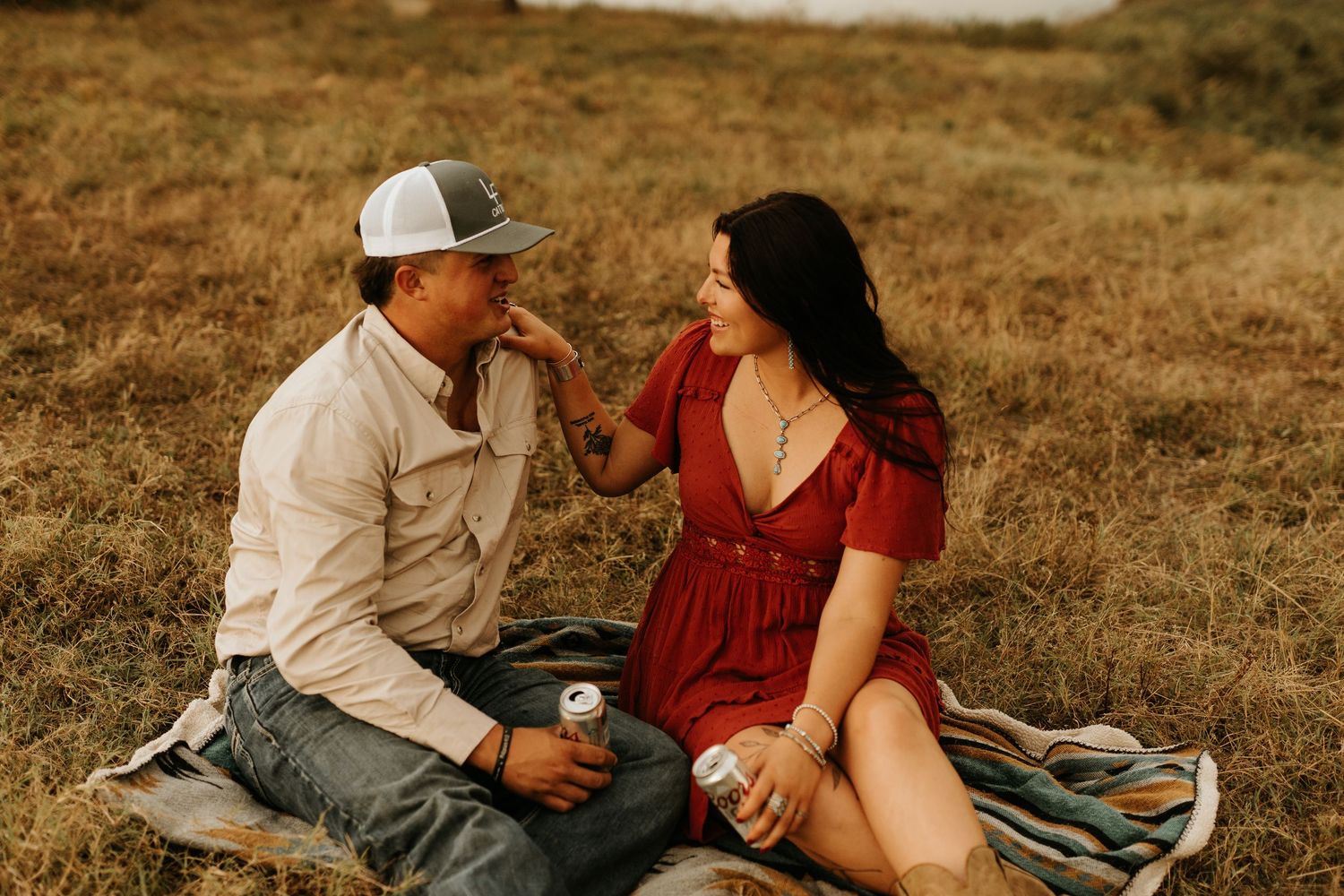 Couple sitting on a blanket in a golden field, sharing a tender moment, woman in red dress touching mans face.