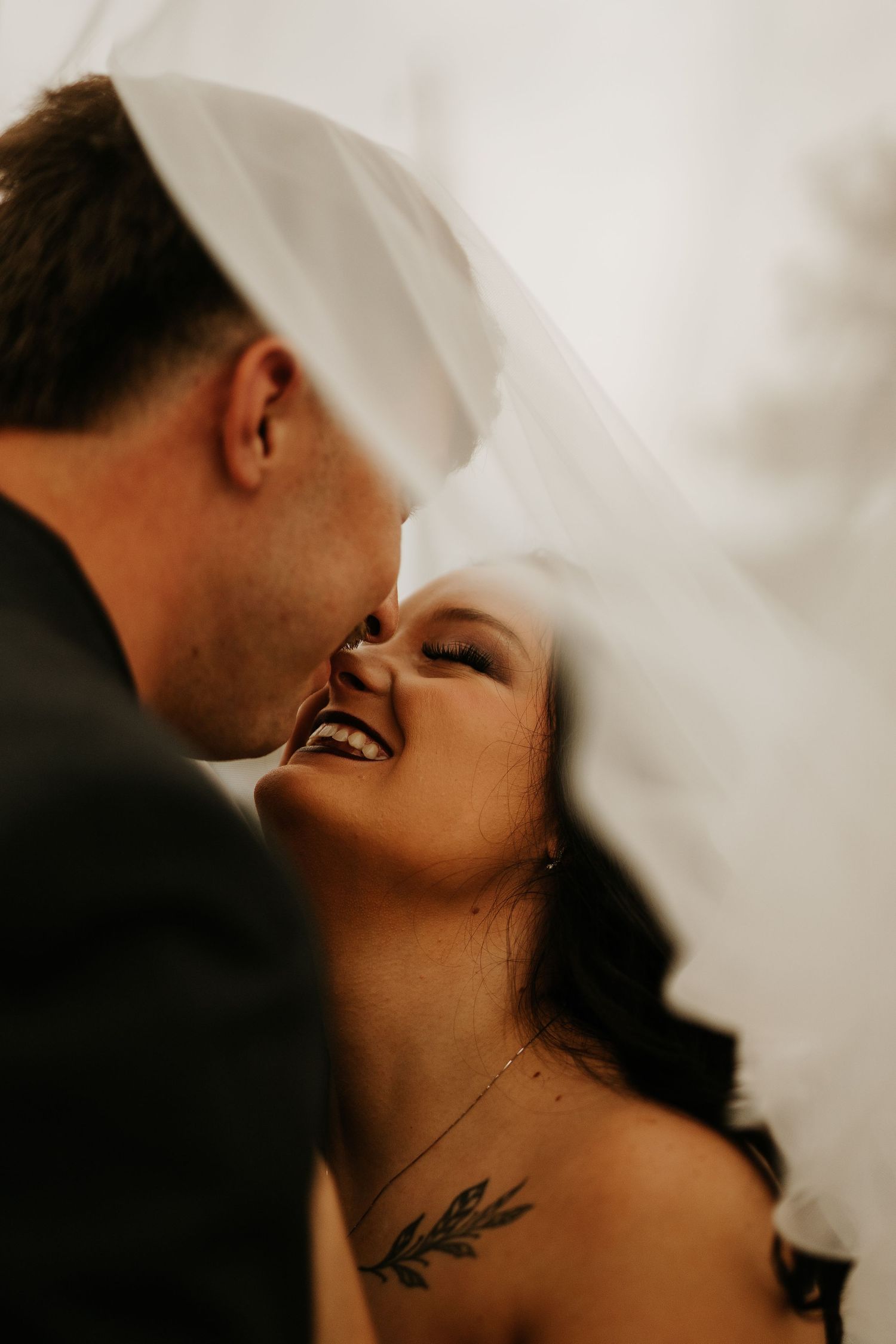 A joyful bride laughs under her veil as the groom leans in close during a romantic wedding portrait.
