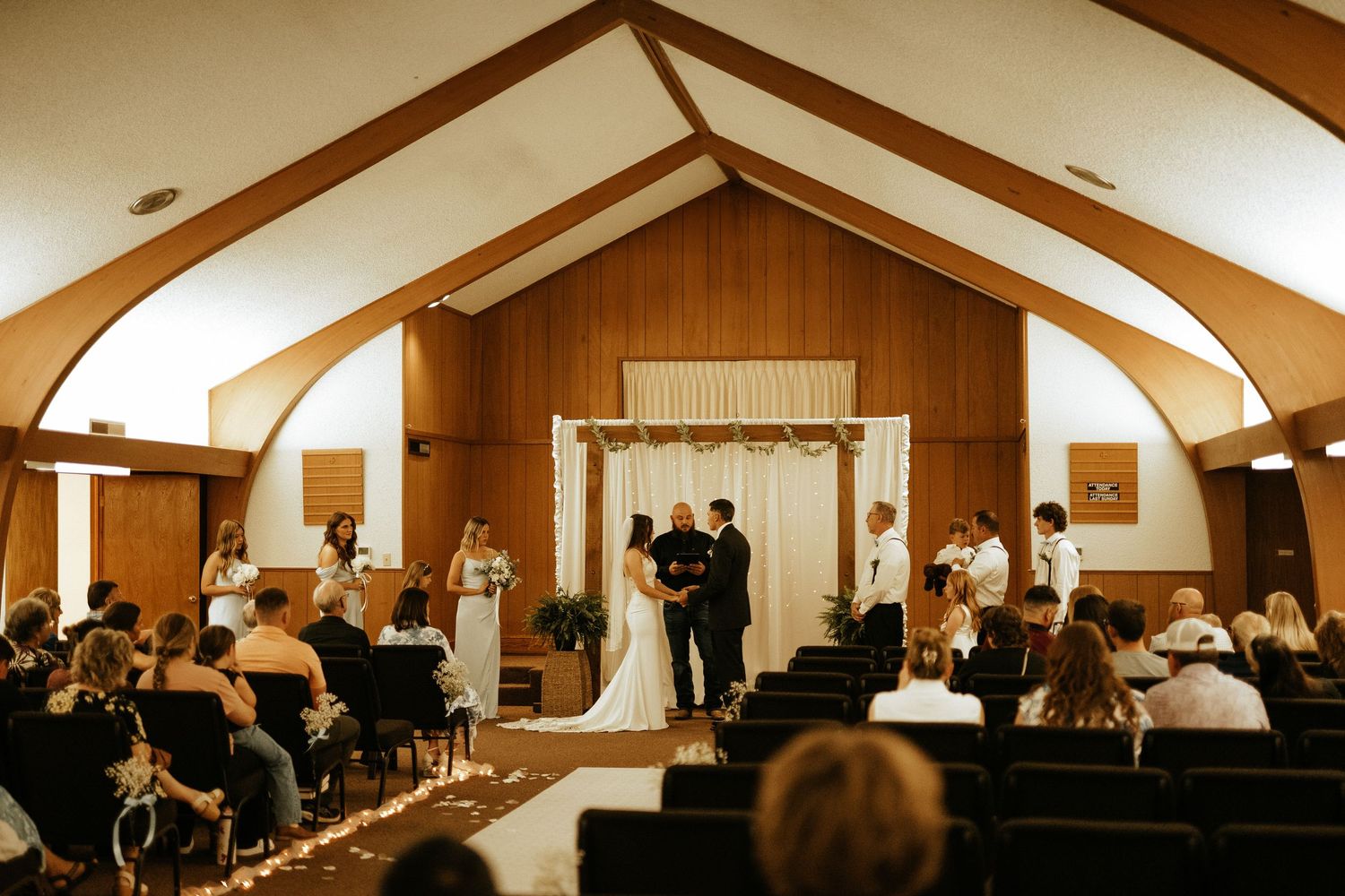 Elegant wedding ceremony in a wood-paneled chapel with vaulted ceilings, couple at altar with floral arch and seated guests.