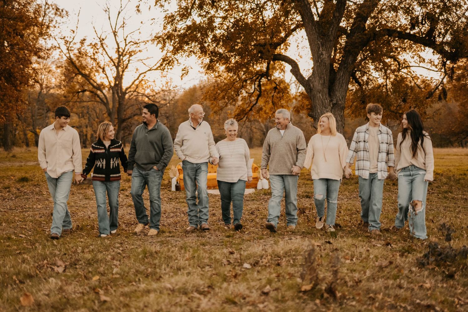 A multigenerational family of nine walks hand-in-hand through a golden autumn park with large oak trees.