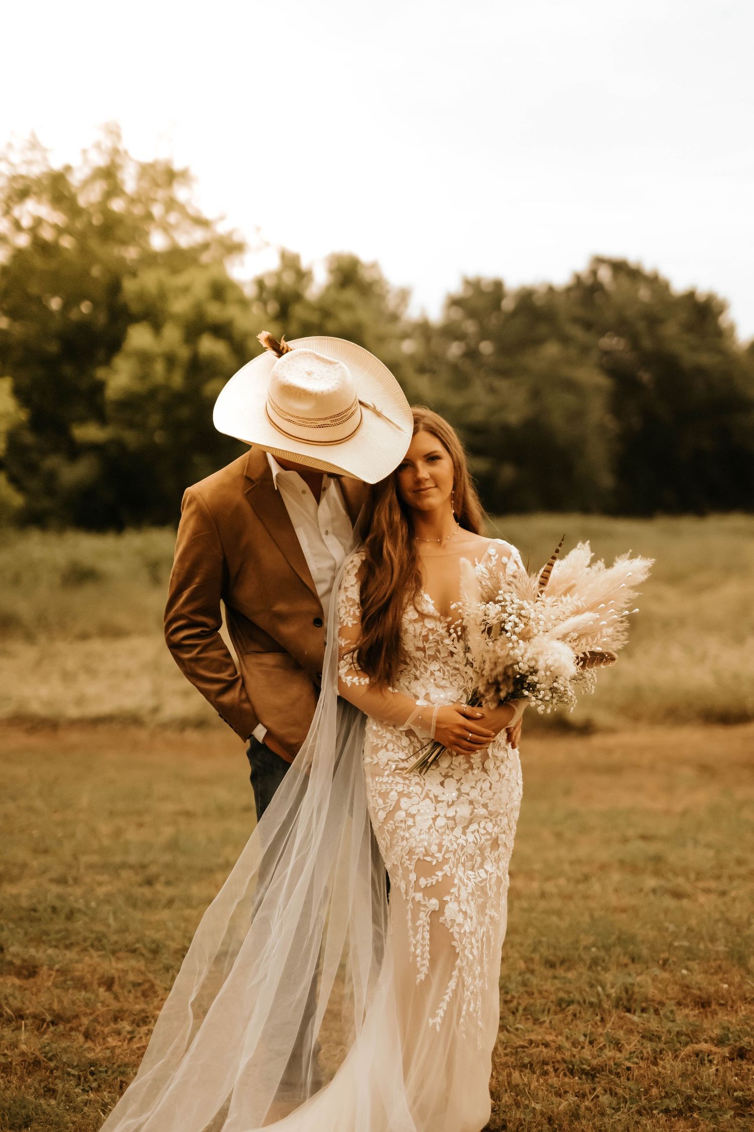 Rustic wedding couple embraces in field, bride in lace dress and groom wearing brown jacket and cowboy hat.