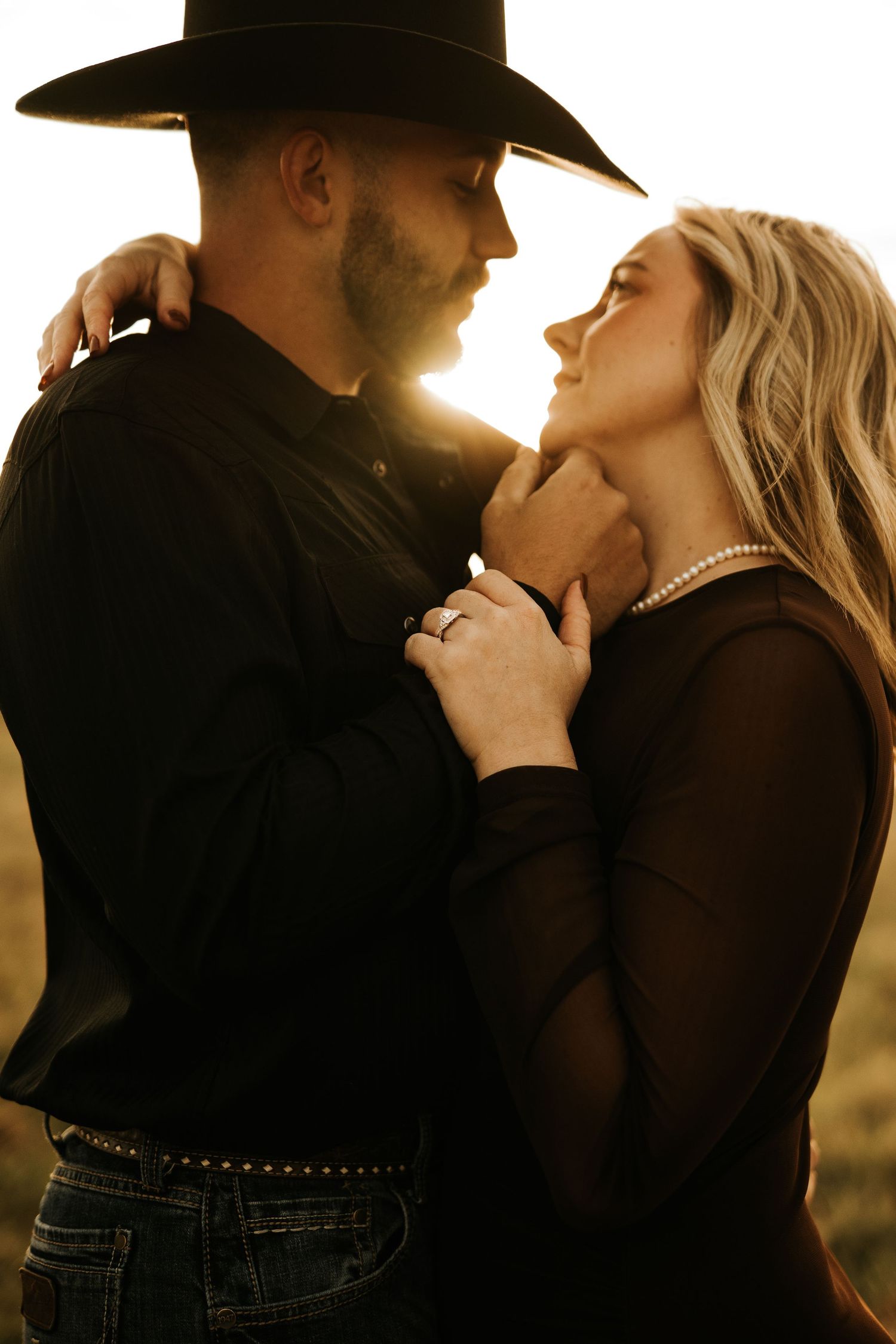 A couple in black western attire shares a romantic golden hour moment, man in cowboy hat gently holding womans face.