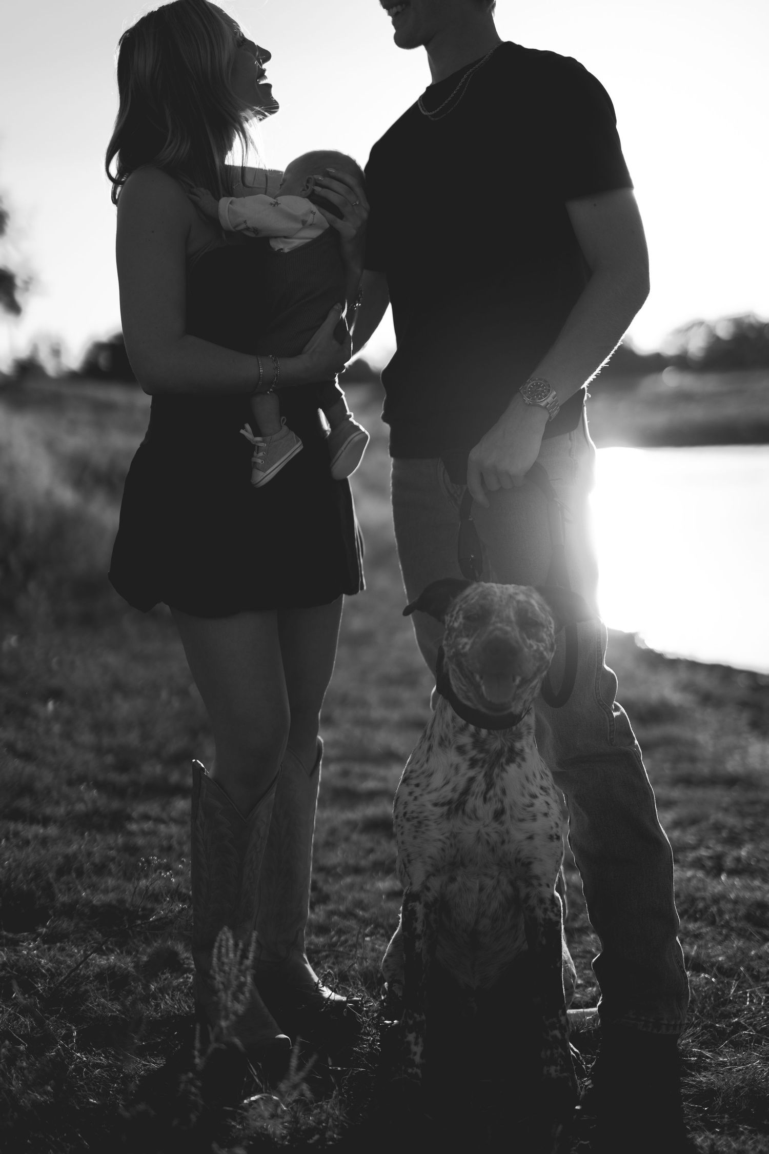Black and white photo of a couple with a baby and spotted dog by a sunlit lake.