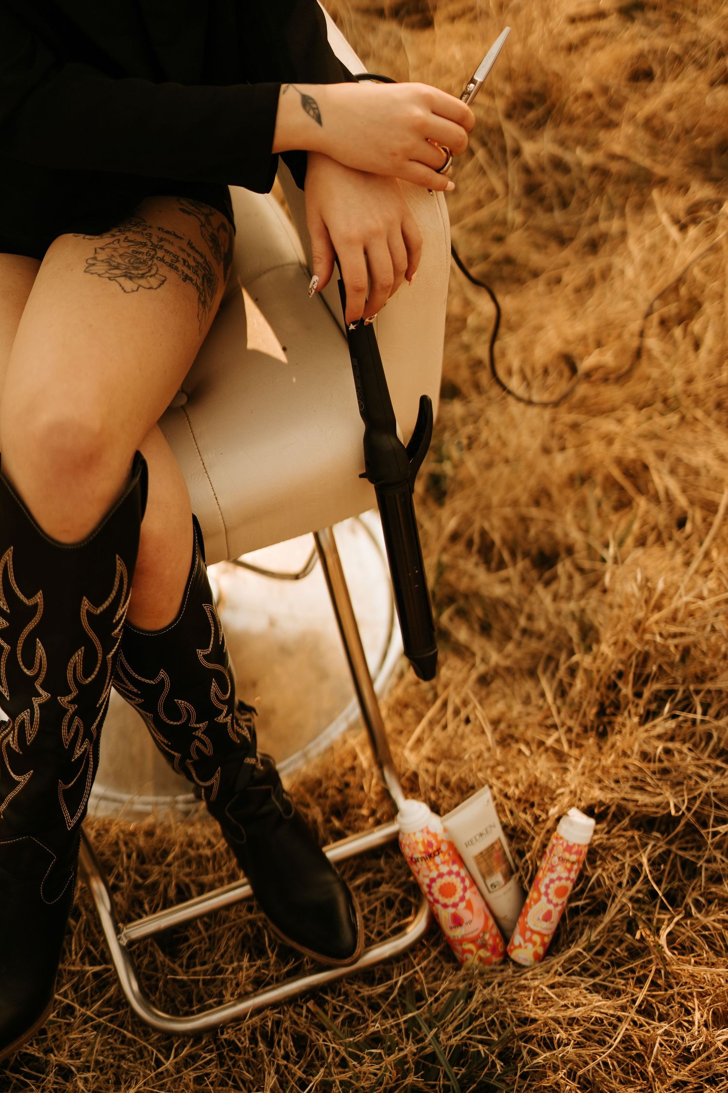 Person with tattoos and cowboy boots sitting on a folding chair in a field with hair tools and products nearby.